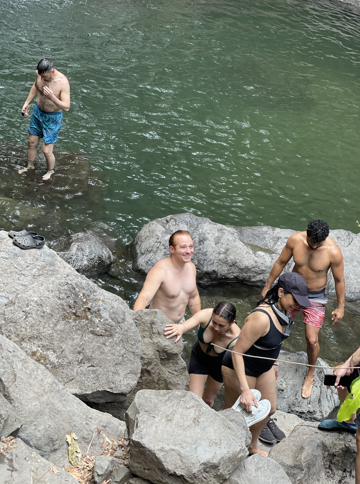 People swimming, sitting, and standing on rocks at a river, with some in swimwear and others in casual clothes.