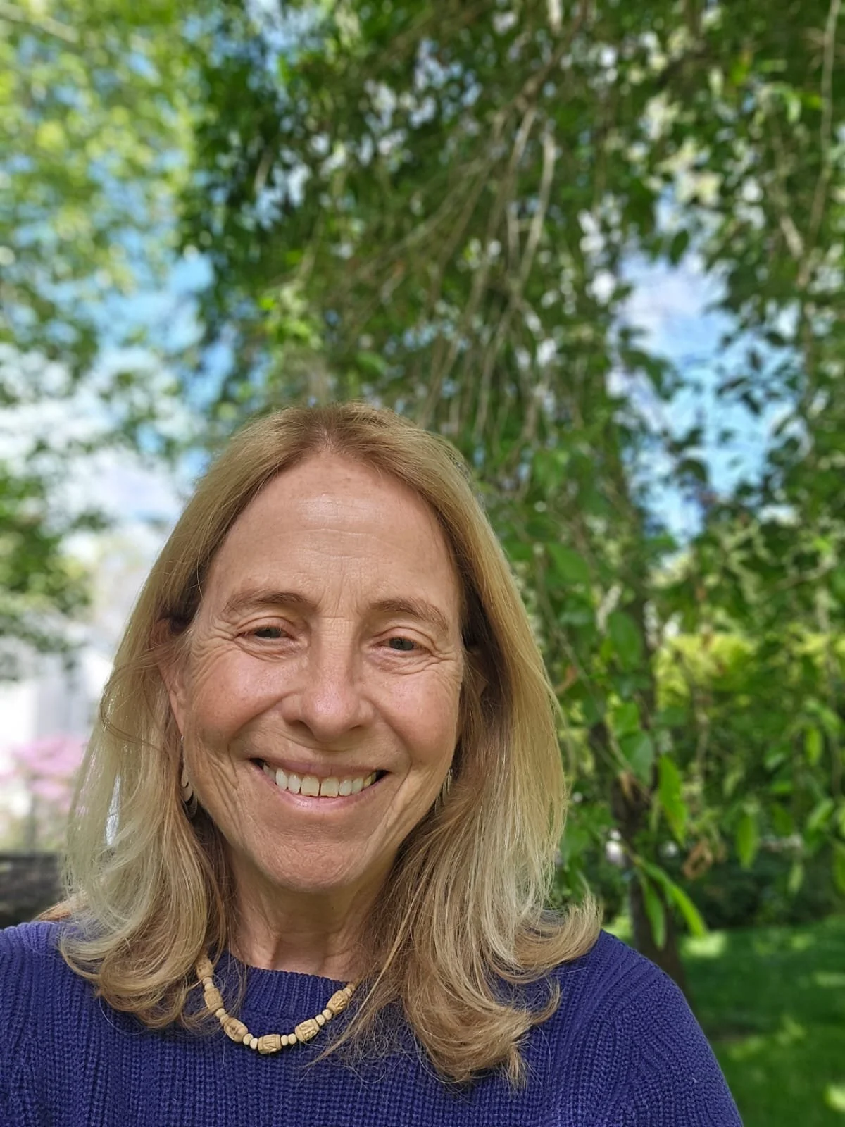 A woman outdoors with a big smile, blonde hair, wearing a blue sweater and a beaded necklace, standing in front of green trees under a partly cloudy sky.