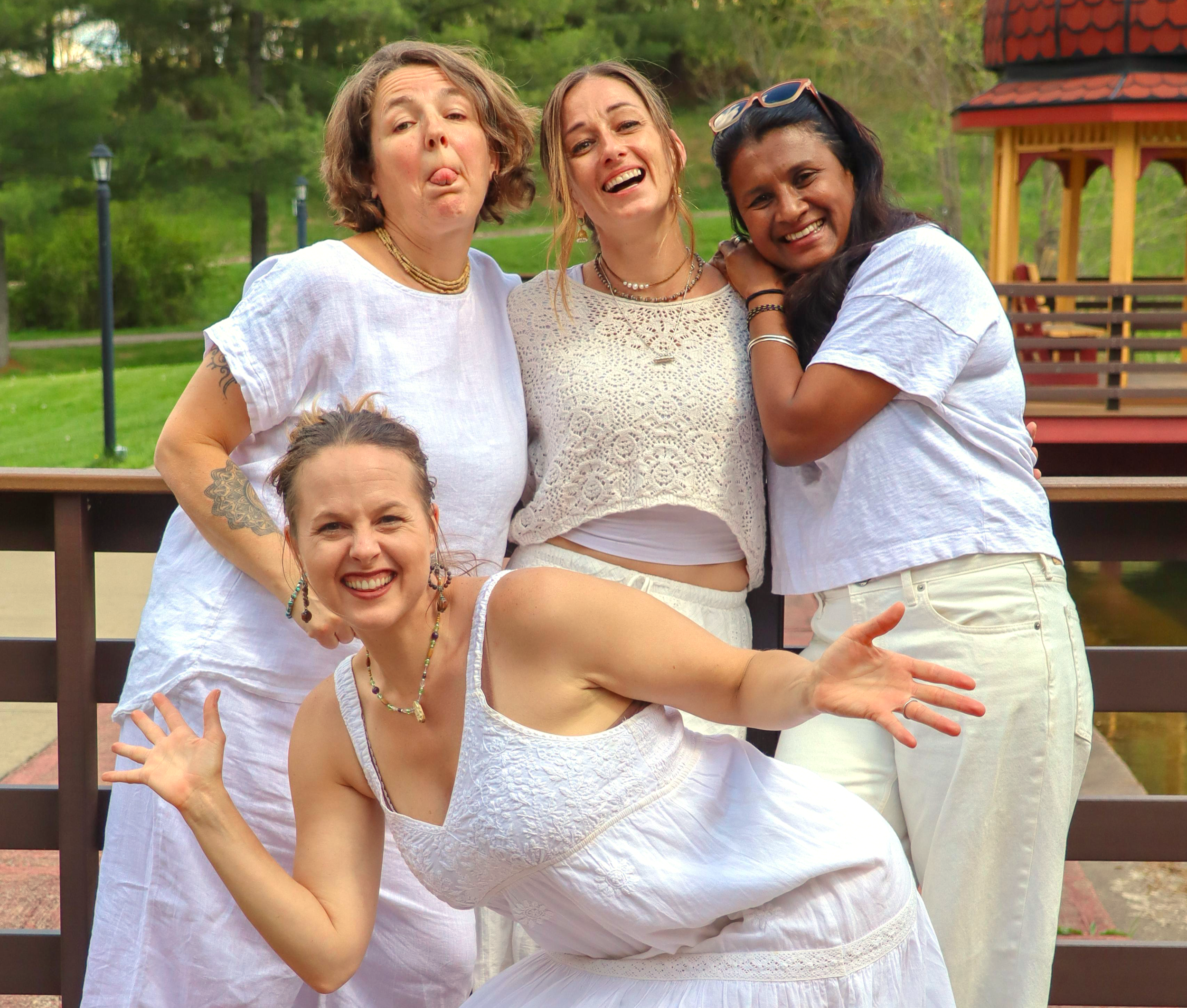 Three women enjoying an outdoor gathering, making silly faces and smiling, with a wooden gazebo and green trees in the background.