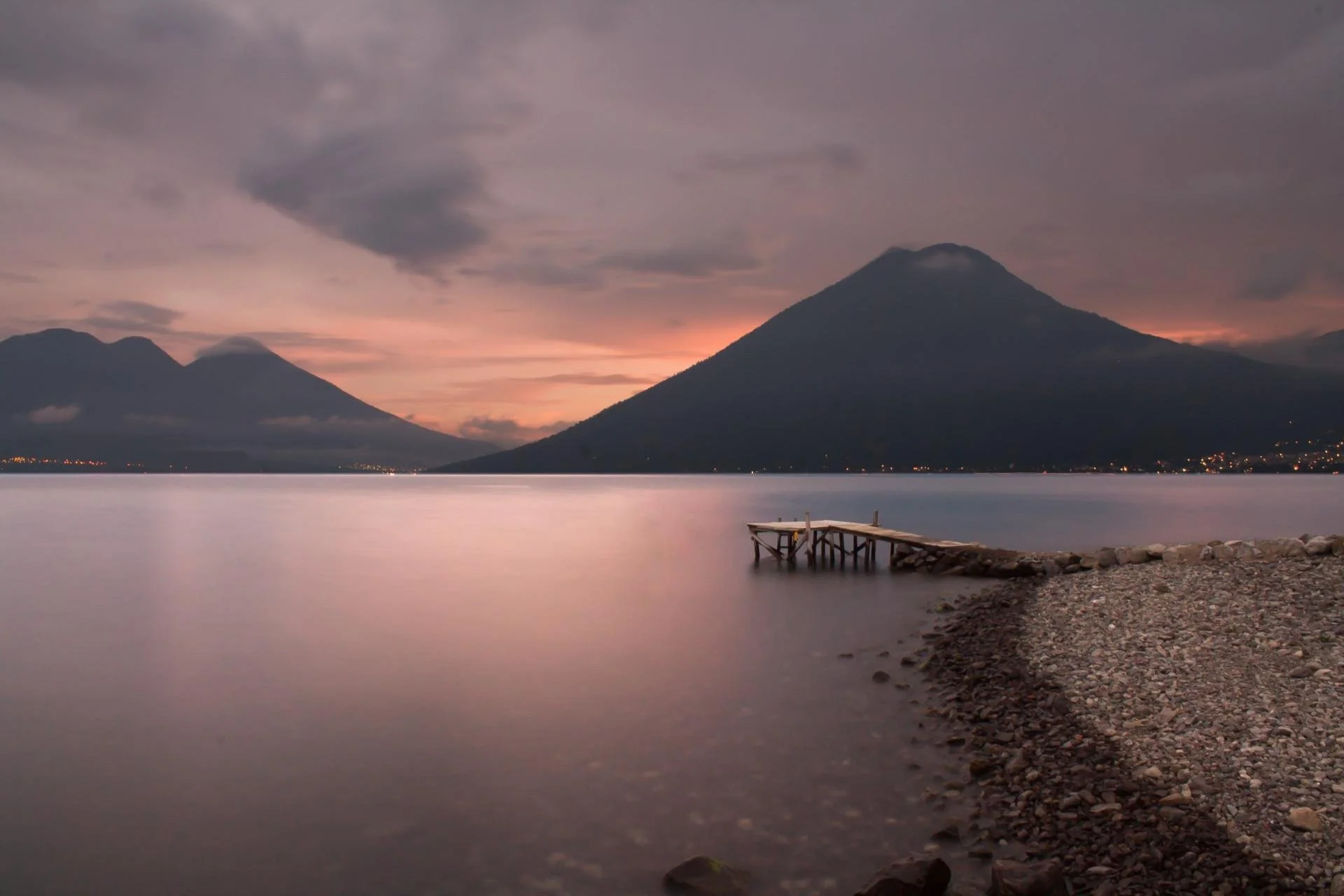 A serene lakeside scene at dusk with two mountains in the background, a wooden pier extending into calm water, and a pebble beach in the foreground.