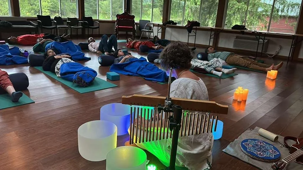 A group of people are lying on yoga mats in a room with large windows, participating in a relaxation or meditation session led by a woman playing a musical instrument, with soft lighting and colorful illuminated bowls nearby.