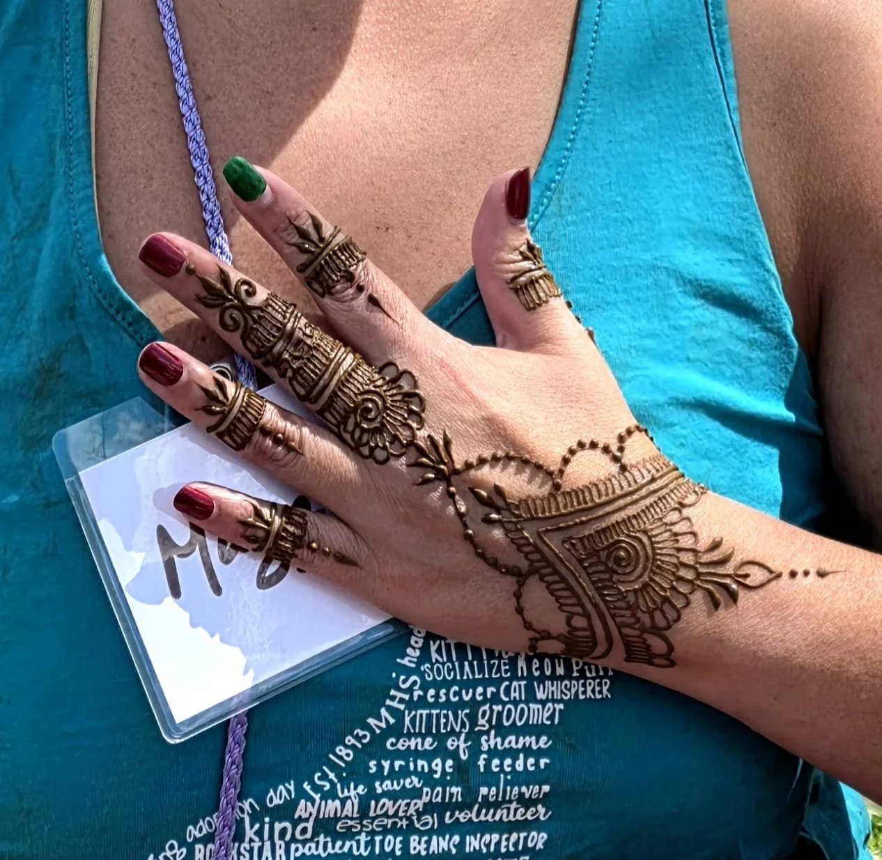A woman's hand with intricate henna designs, wearing multiple rings and nail polish in red and green, resting on a person's chest. The person is wearing a turquoise tank top, and there is a laminated name badge attached to a purple lanyard.