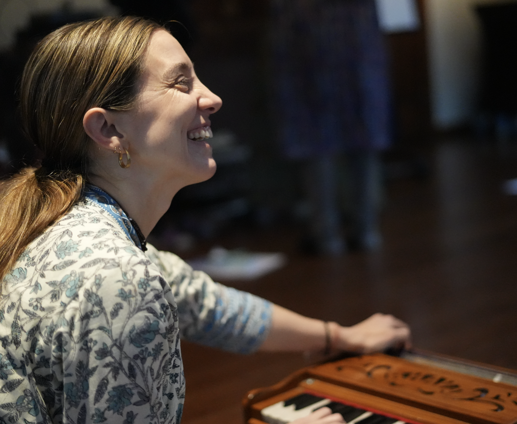 A woman with red hair, wearing earrings and a patterned shirt, playing a piano in a dimly lit room, smiling and looking towards the right side of the image.