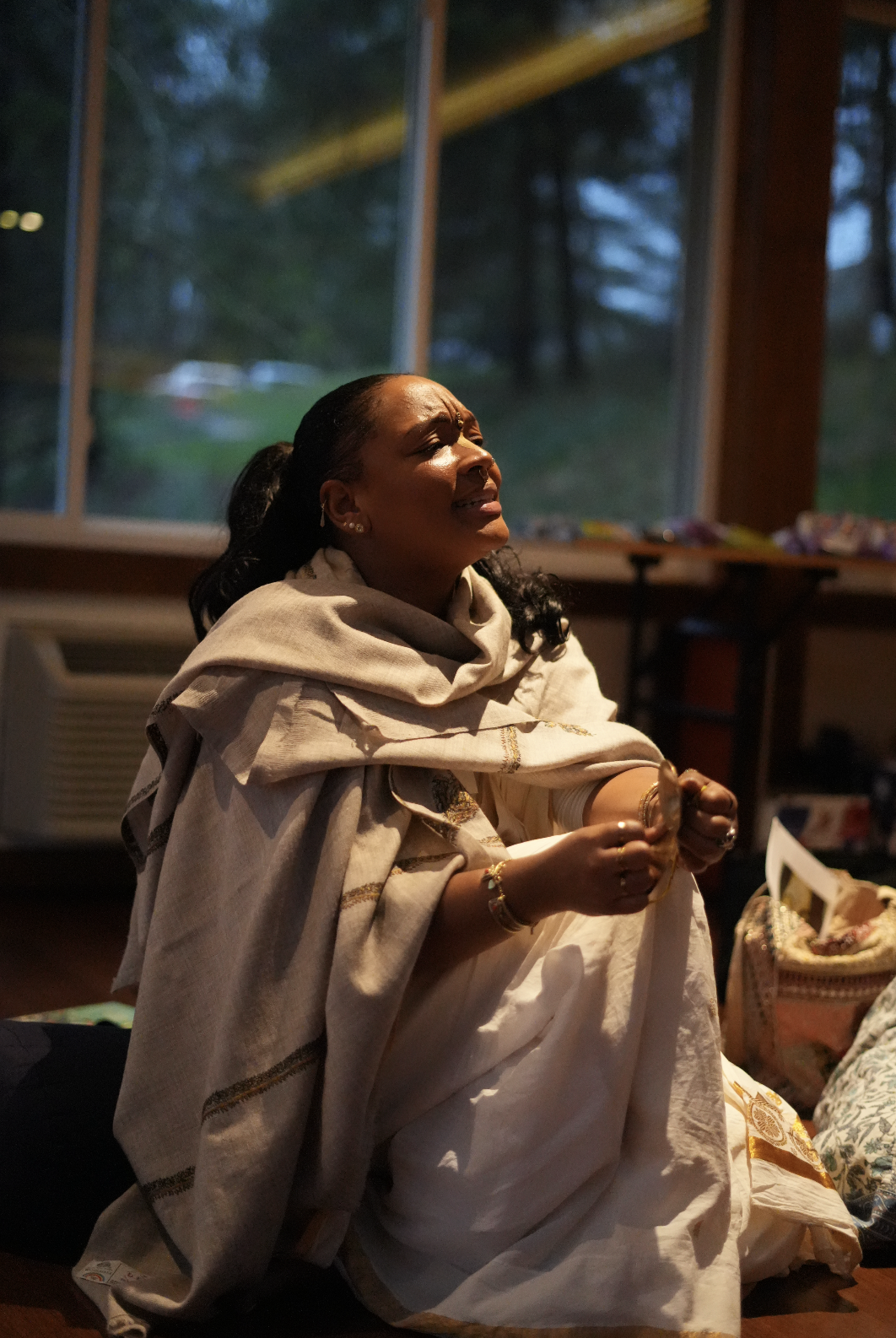 A woman in traditional clothing appears emotional, sitting indoors near a window with a forest background, possibly praying or mourning.