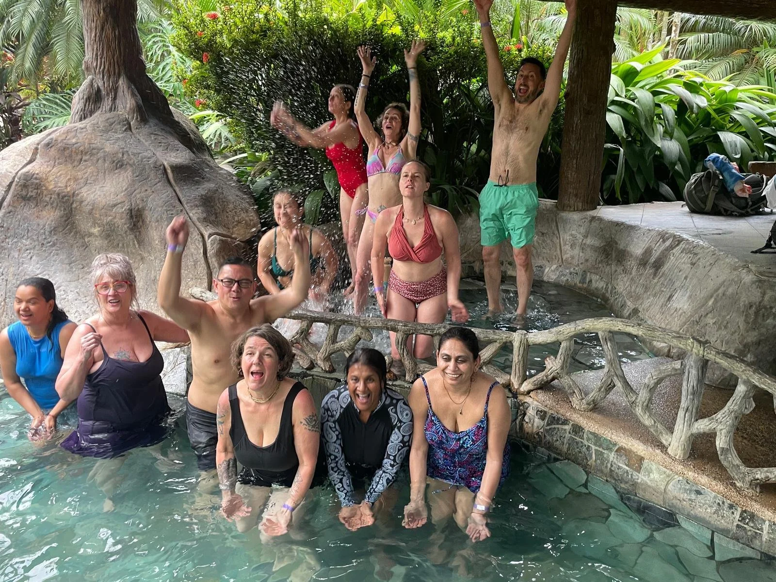 A group of people enjoying and playing in a small pool with a waterfall feature in a lush, tropical setting.
