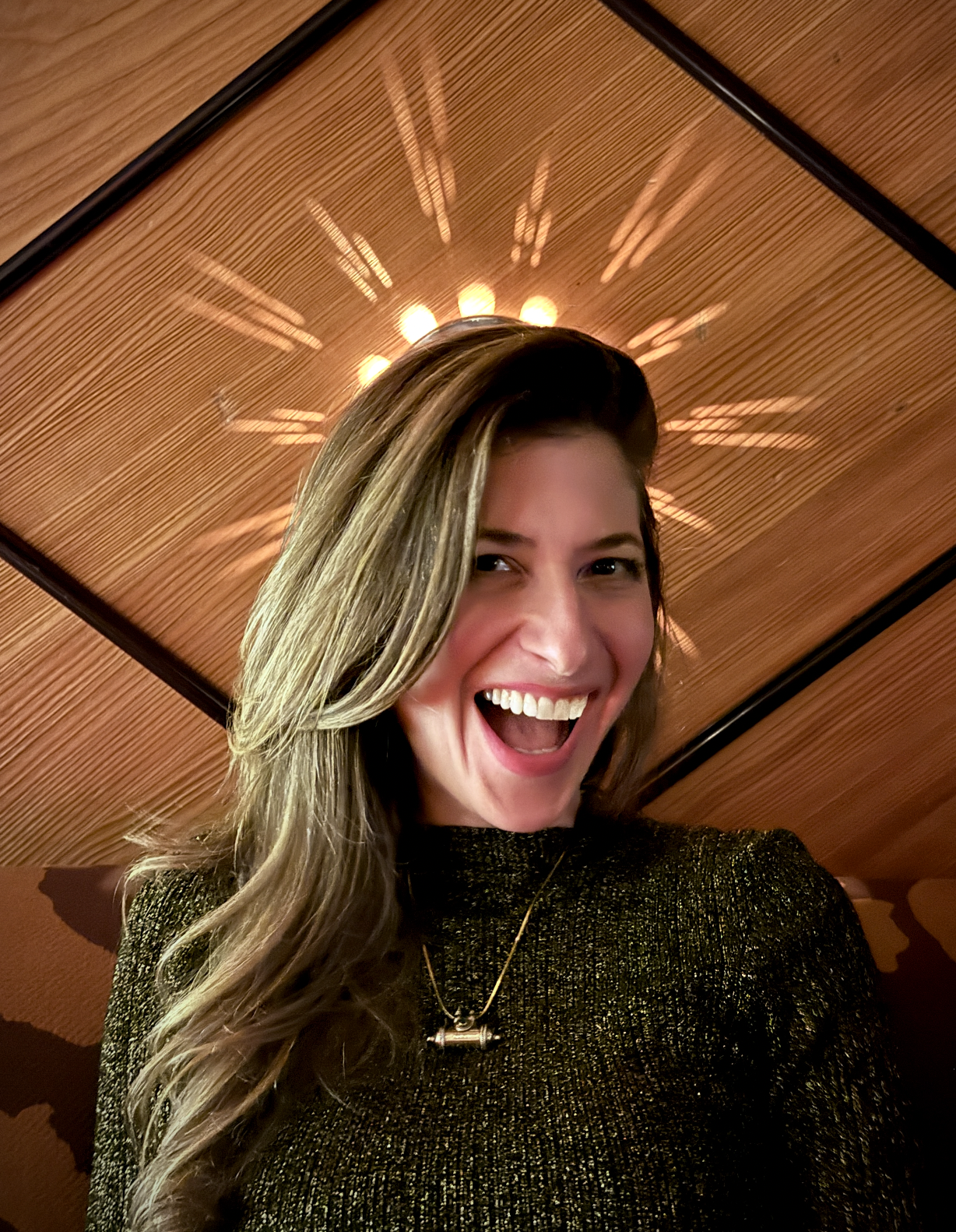 A woman with long, wavy hair smiling and laughing, wearing a sparkly black top and a necklace, with a circular light fixture on a wooden ceiling in the background.