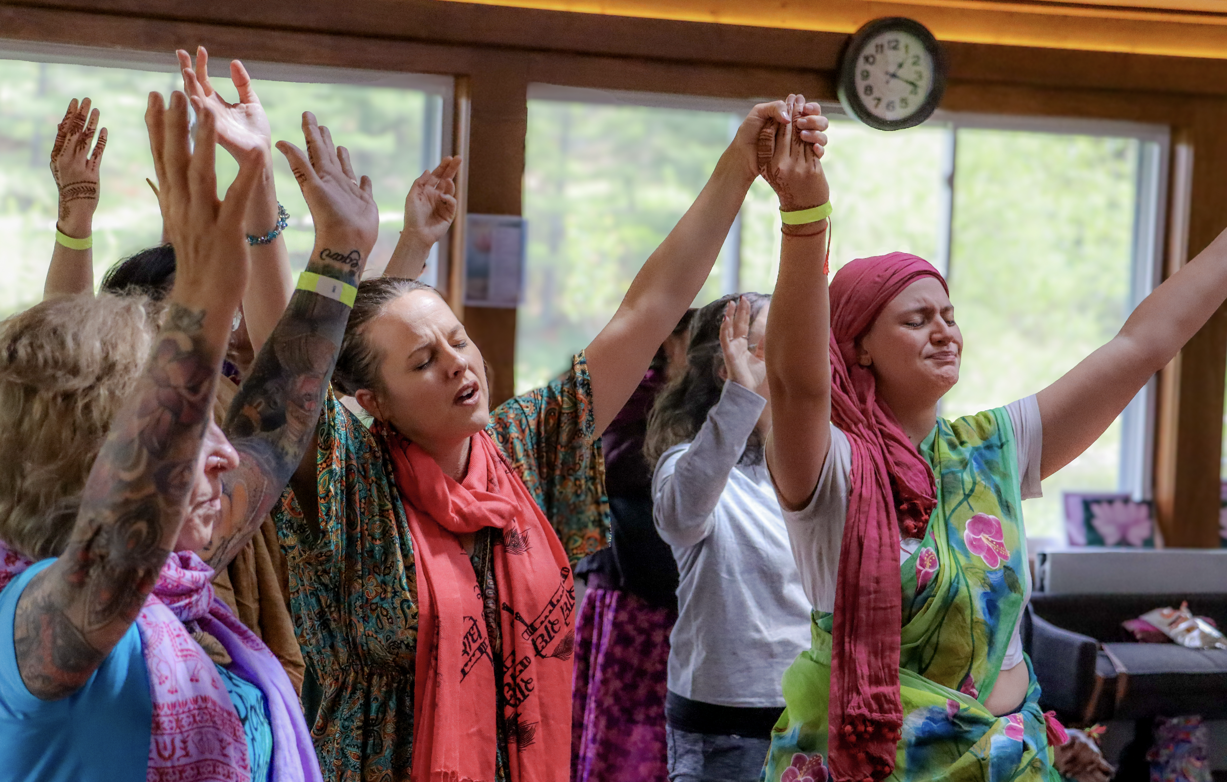 A group of diverse women with tattoos and colorful clothing participating in a meditation or prayer session with their eyes closed and hands raised, inside a wooden-floored room with large windows and a clock on the wall.