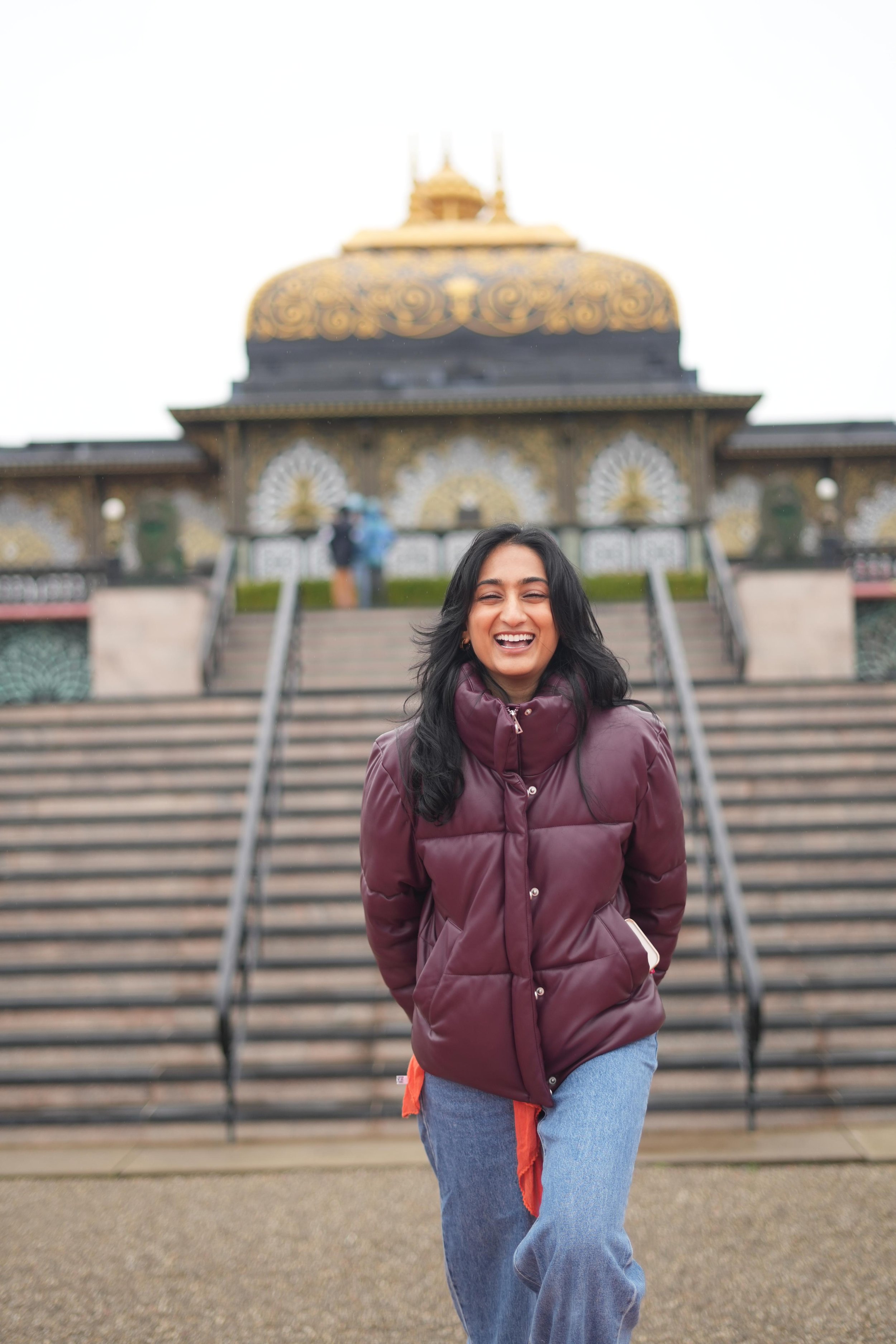 A smiling woman in a maroon puffy jacket and light blue jeans standing on stairs in front of a temple with intricate gold and black decoration, with two people blurred in the background.