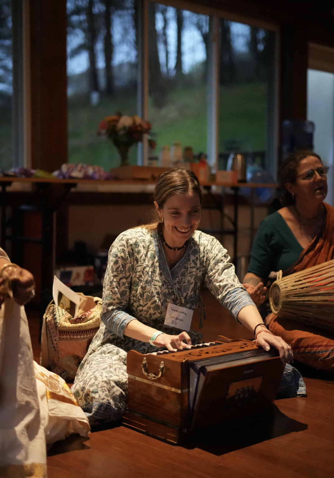 A woman smiling and playing a harmonium at an indoor gathering, with people and a window with trees in the background.