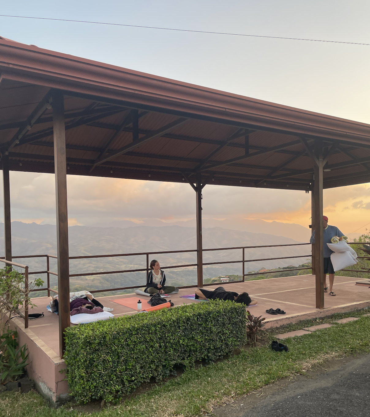 People practicing yoga on a covered outdoor terrace overlooking mountains at sunset, with some lying on mats and one woman sitting cross-legged.
