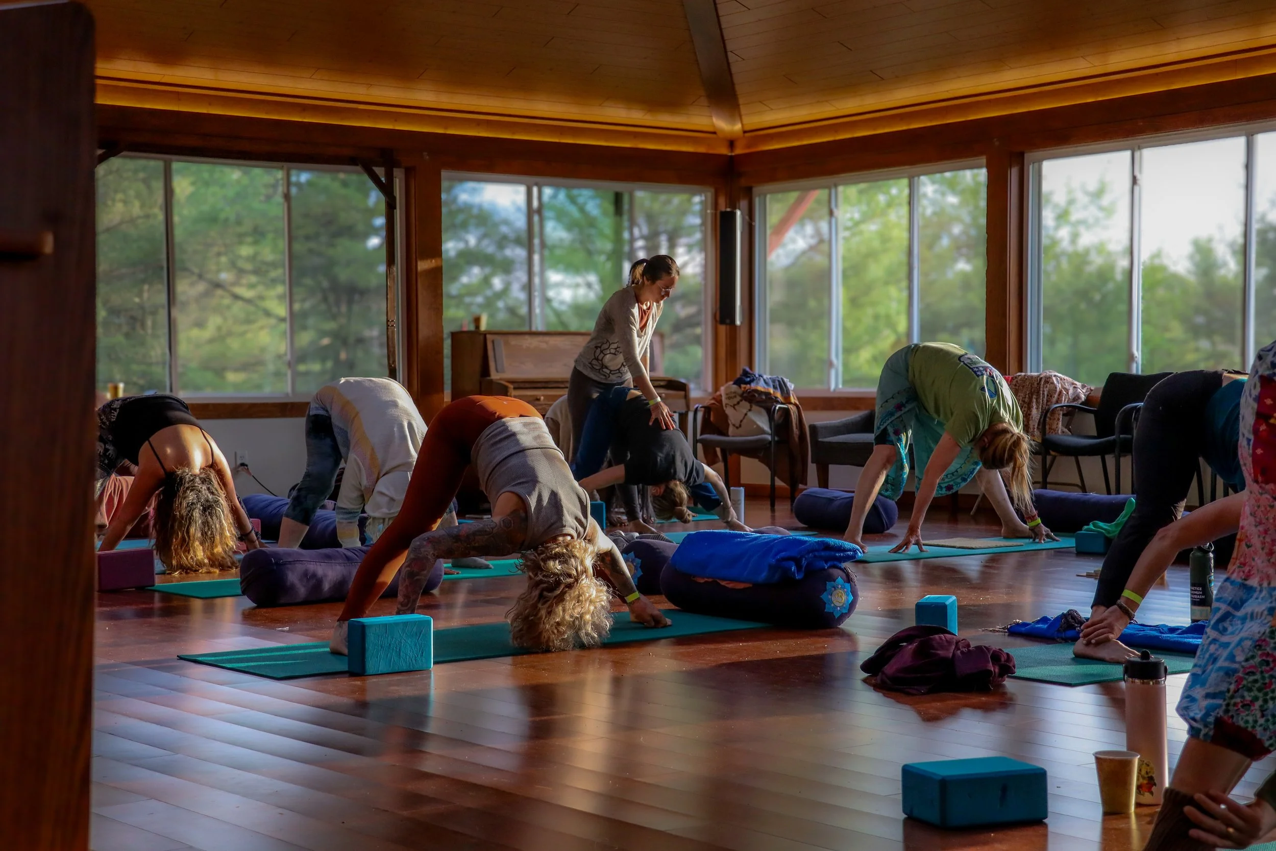 A group of people practicing yoga in a spacious, sunlit room with large windows and wooden walls, performing downward dog poses.