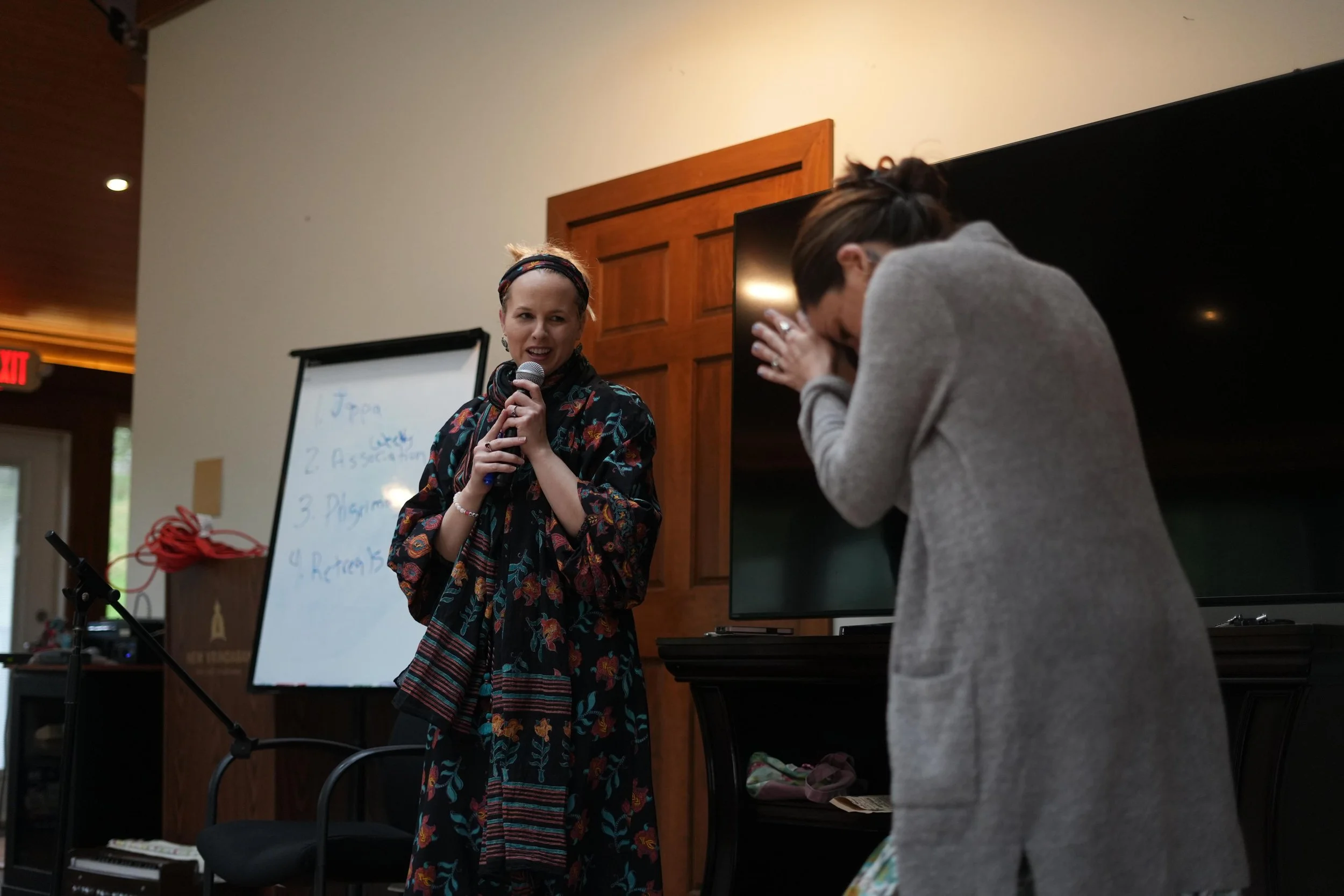 A woman with a floral dress and a scarf speaking into a microphone during a presentation or workshop, with another woman in a gray sweater covering her face in front of her, in a room with wooden paneling and a whiteboard.