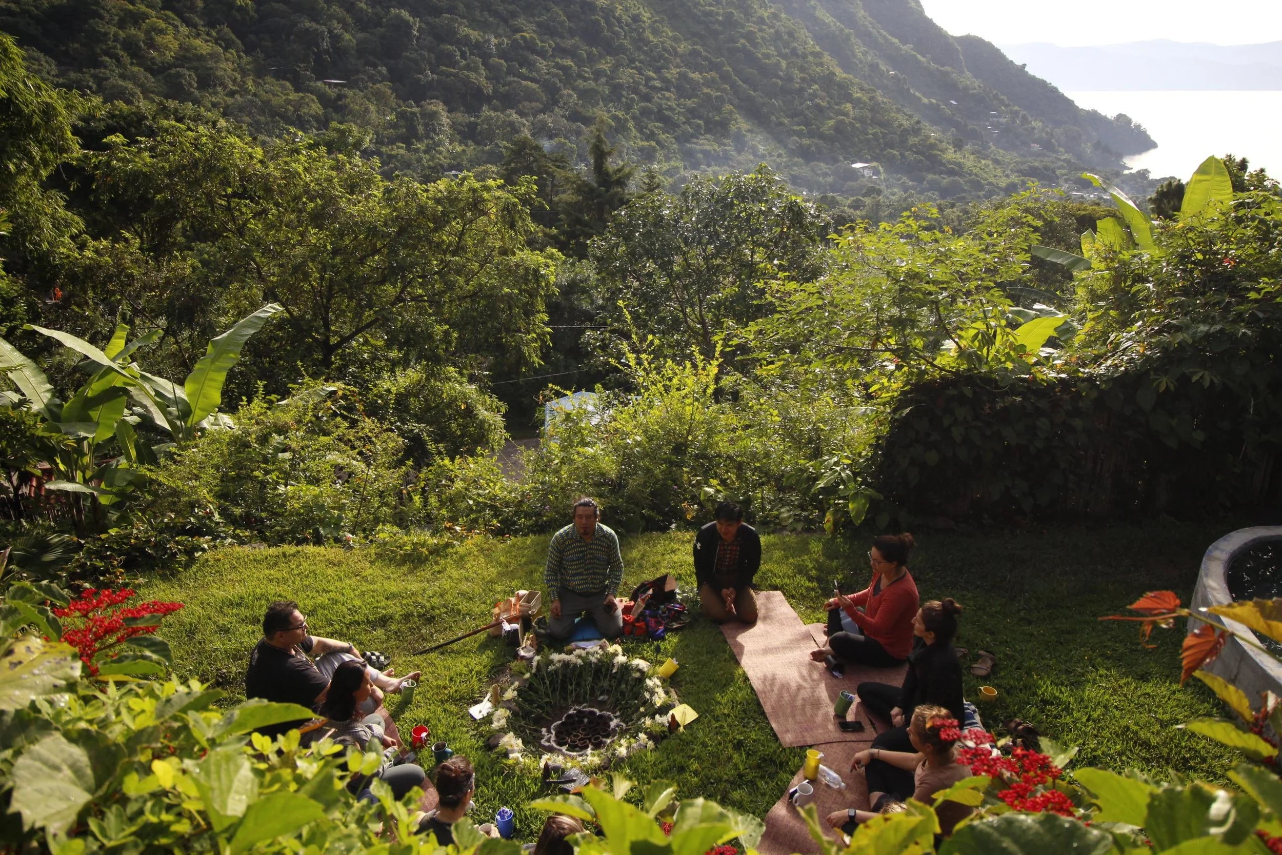 Group of people sitting on the grass in a circle during a meditation session with lush green forest and mountains in the background.