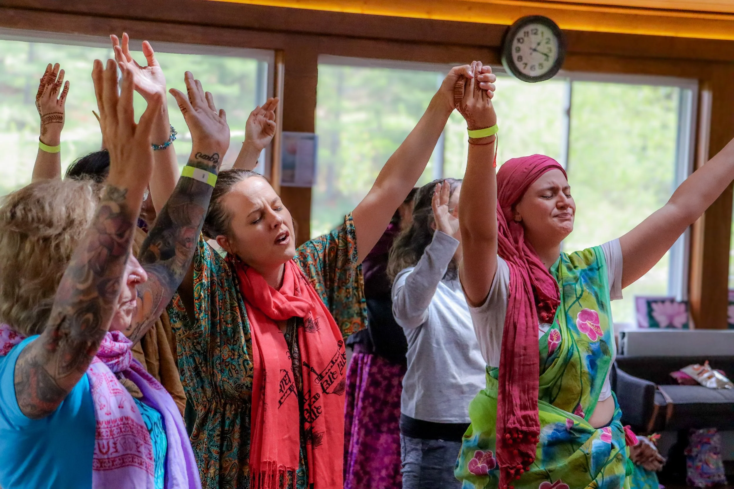 A group of people with tattoos and colorful clothing praying with raised hands in a bright room with large windows and a clock.