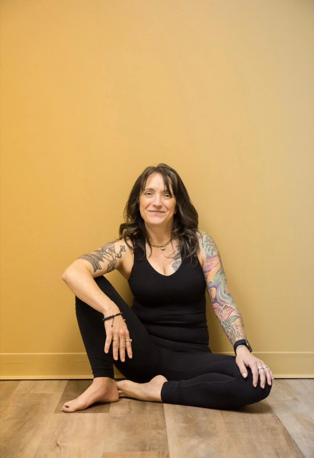 A woman sitting on the floor against a yellow wall, wearing a black tank top and black leggings, with tattoos on both arms, smiling at the camera.