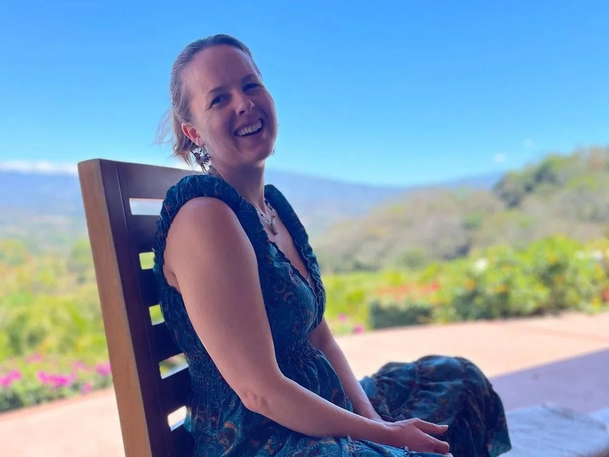 A smiling woman with short blonde hair sitting on a wooden chair outdoors, with a scenic landscape of green hills and a blue sky in the background.