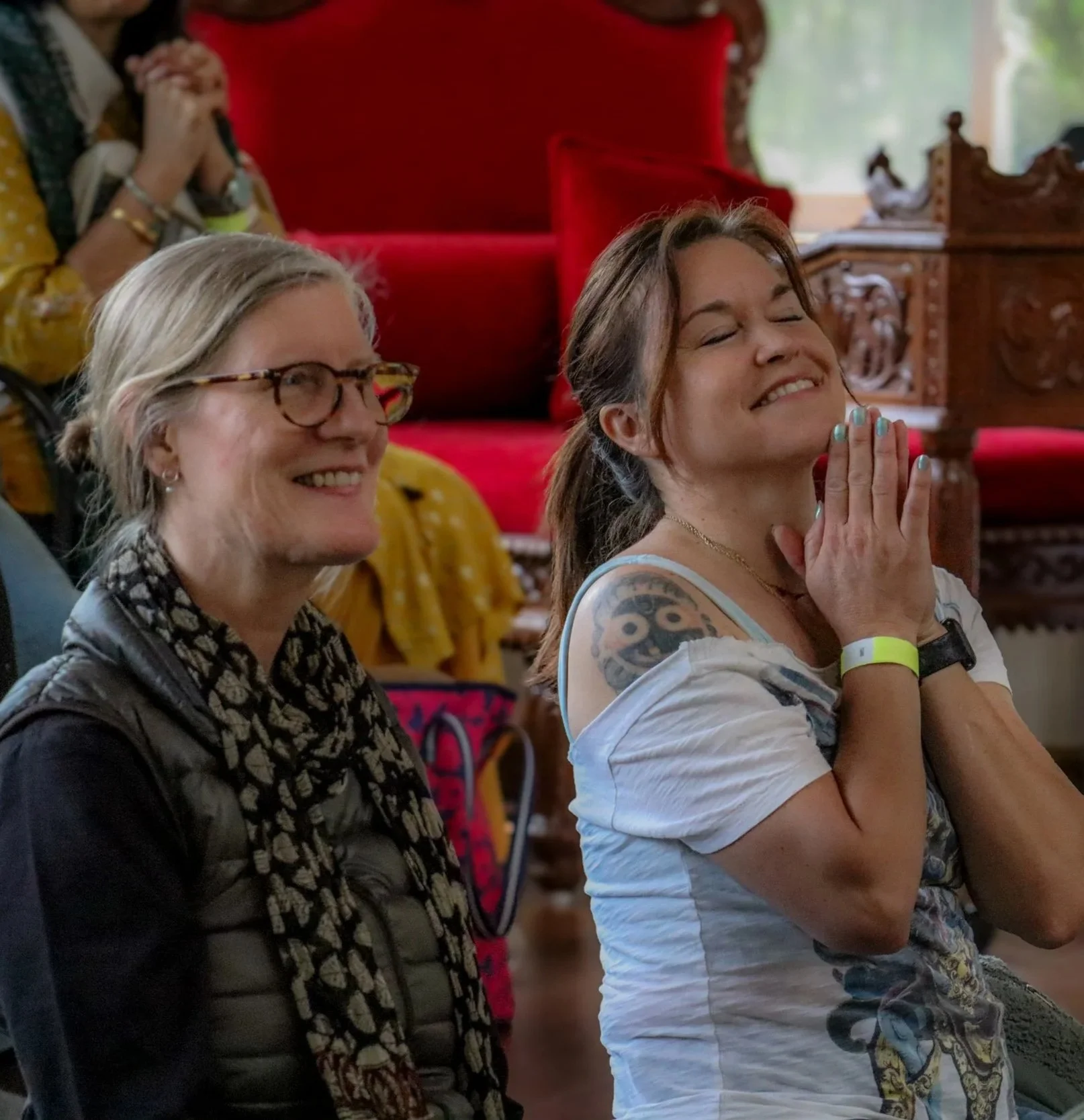 Two women sitting and smiling during an indoor event, one with glasses and the other with closed eyes and hands pressed together in a prayer-like pose, with a red cushioned antique-style chair and wooden furniture in the background.