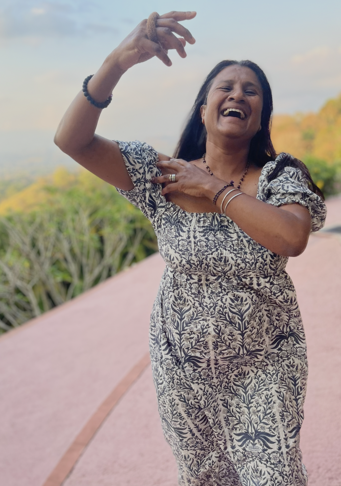 A smiling woman with long dark hair wearing a patterned dress, standing outdoors on a pink pathway with greenery and distant trees in the background.