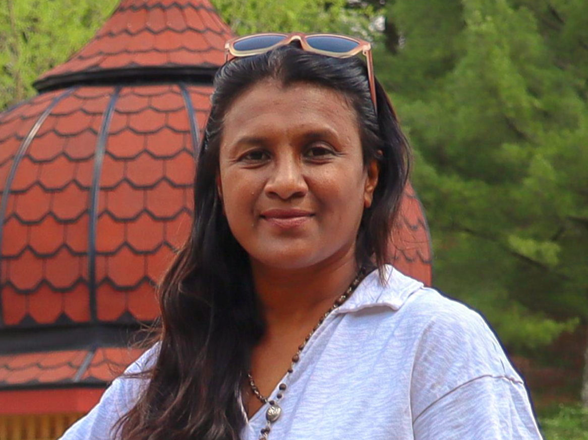 A woman with long dark hair, sunglasses on her head, wearing a white shirt and a beaded necklace, standing outdoors in front of a red-roofed structure and green trees.
