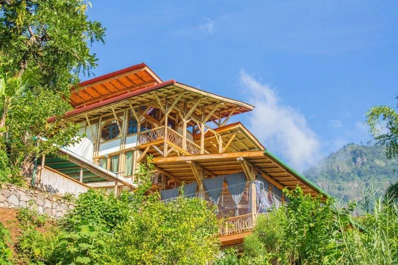 Multi-level wooden house built on a hillside surrounded by green trees and mountains, with a blue sky above.