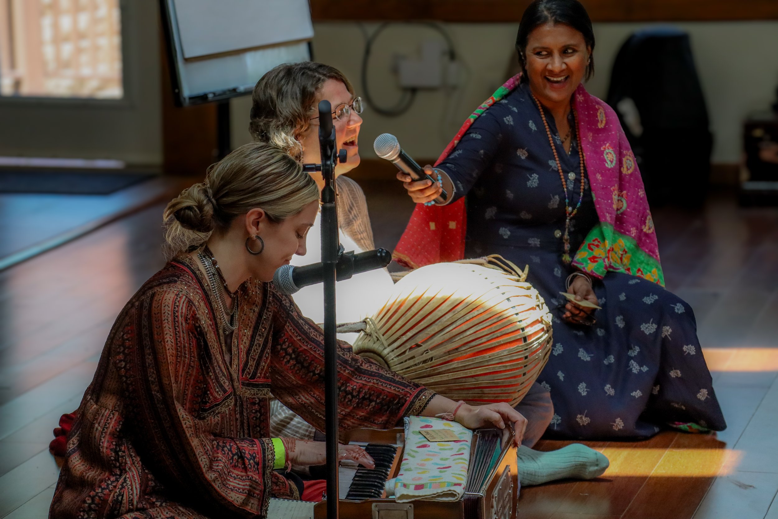 Three women are performing traditional Indian music, with two playing harmoniums, one playing tabla, and one woman singing into a microphone, while seated on the floor.
