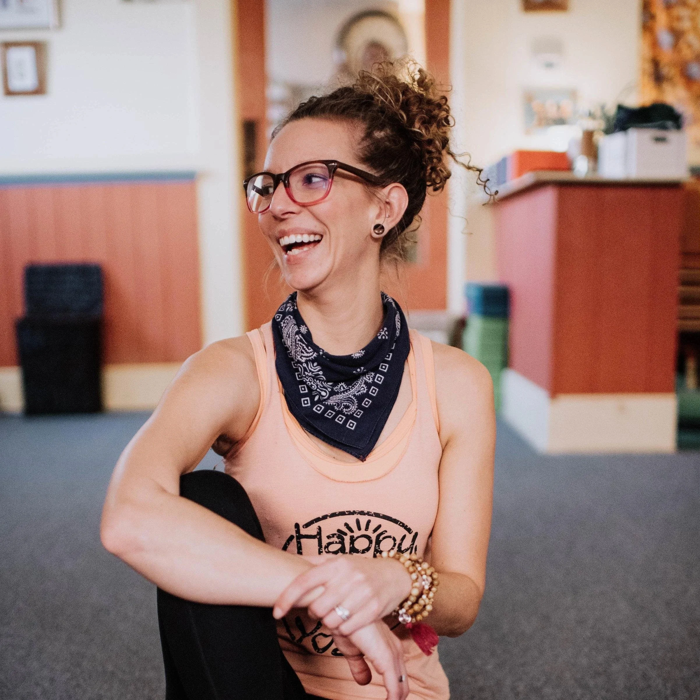 A woman with curly hair, wearing glasses, a bandana, and a peach-colored tank top, smiling and holding a small pink object.