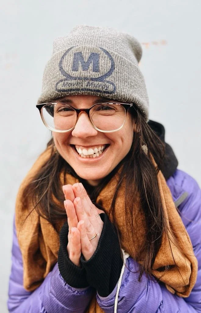 Smiling woman wearing glasses, a gray beanie with a logo, a purple jacket, and a brown scarf, with her hands pressed together in front of her chest.