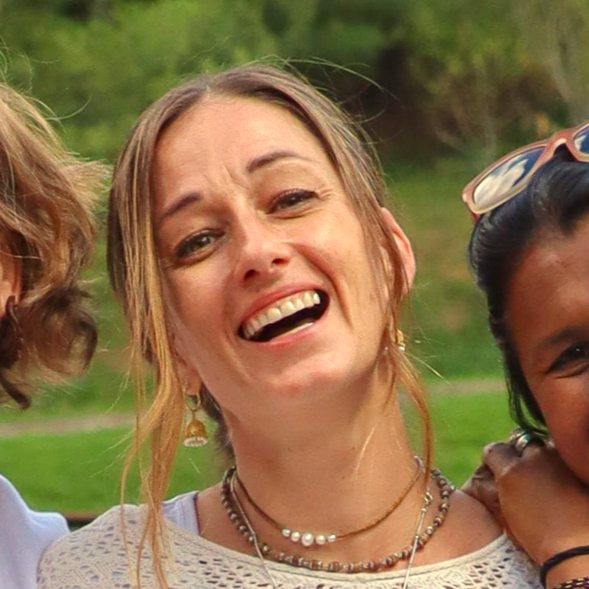 A woman smiling outdoors with green trees in the background, wearing necklaces and earrings.