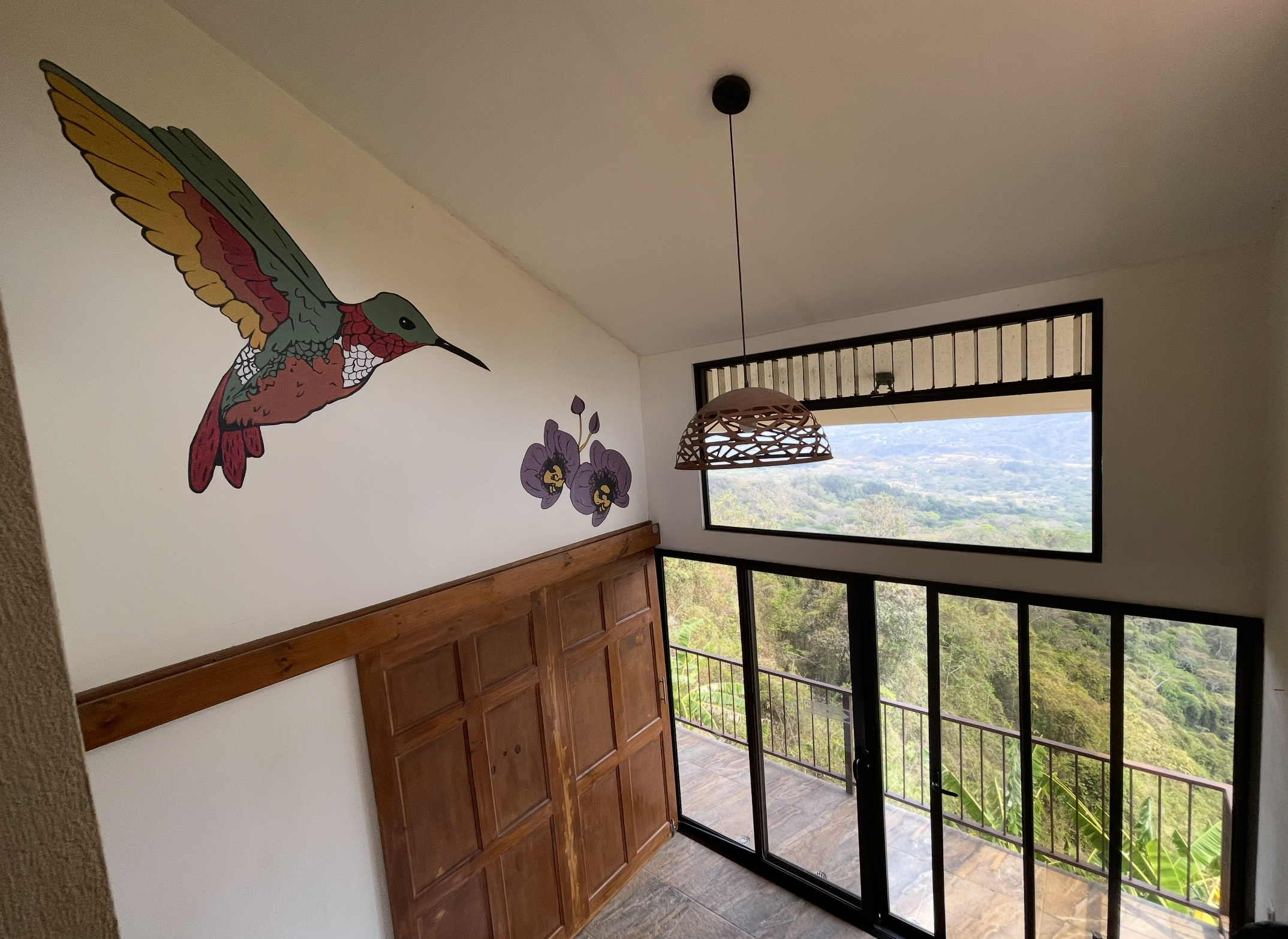 Interior of a room with a wooden cabinet, a colorful hummingbird mural on the wall, purple flowers, a hanging light fixture, window with a view of lush green landscape and hills outside.