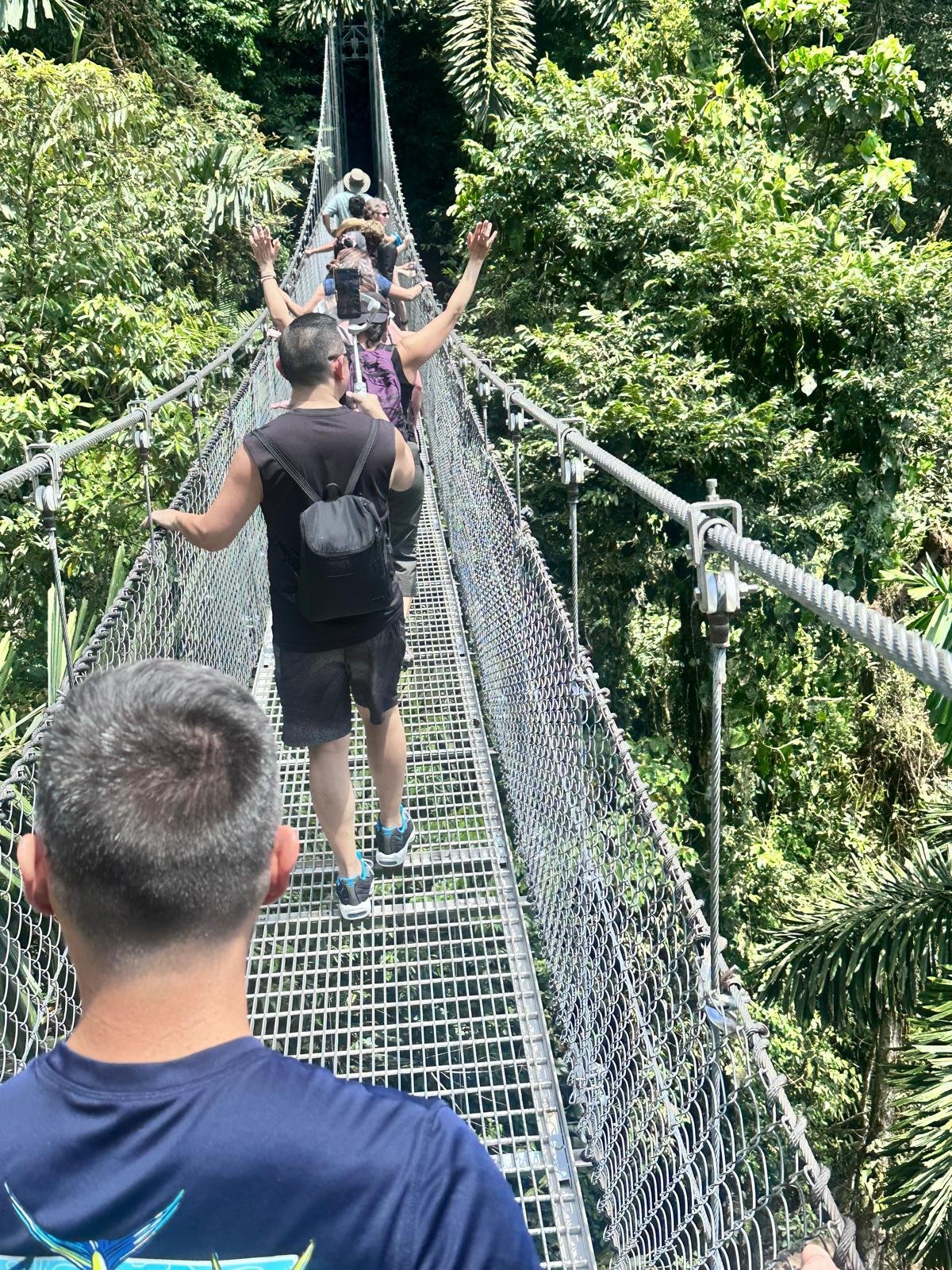 Tourists walking on a narrow suspension bridge made of metal grating high above a lush jungle canopy.