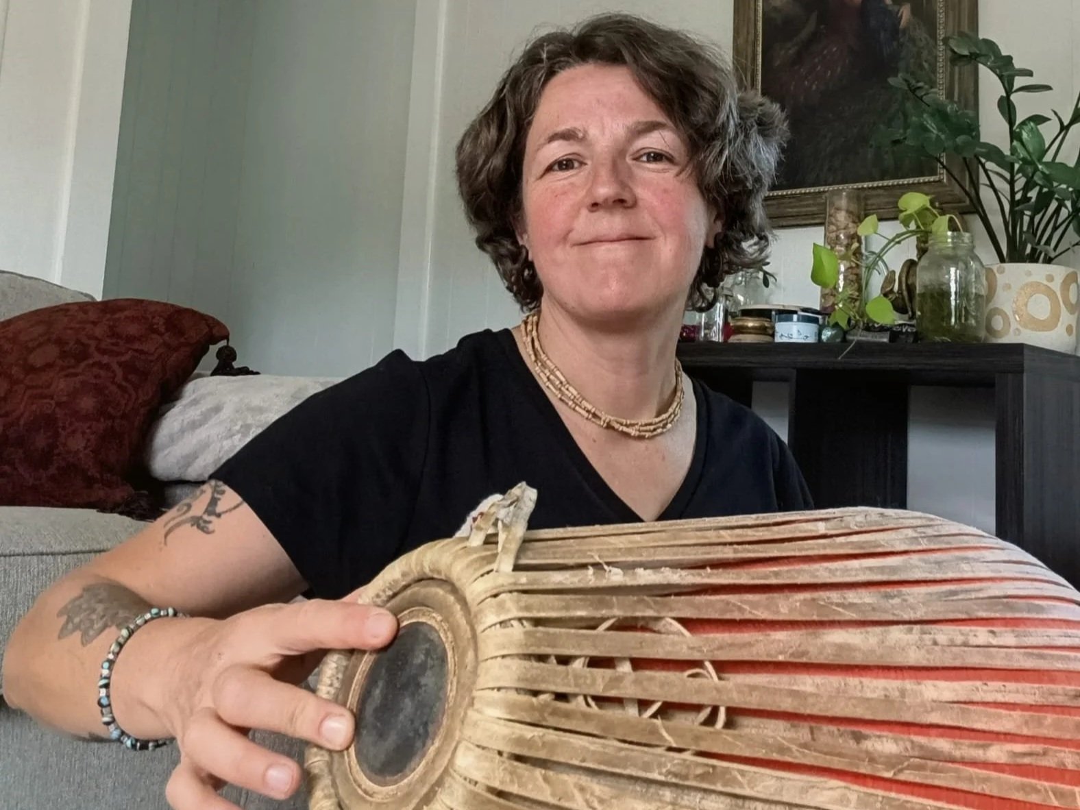 A woman with curly short hair, wearing a black shirt, jewelry, and tattoos, sits on a couch indoors holding a large, wrapped, fish-shaped object made of wooden slats.