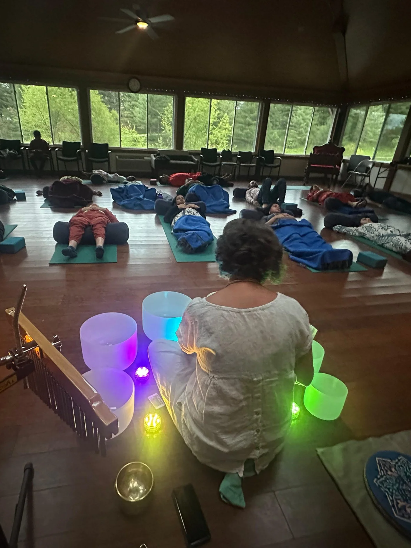 A group of people lying on yoga mats in a circle in a spacious room with large windows overlooking green trees, with a light display of colorful glowing bowls and a woman sitting in front of the circle, possibly leading a meditation or relaxation session.