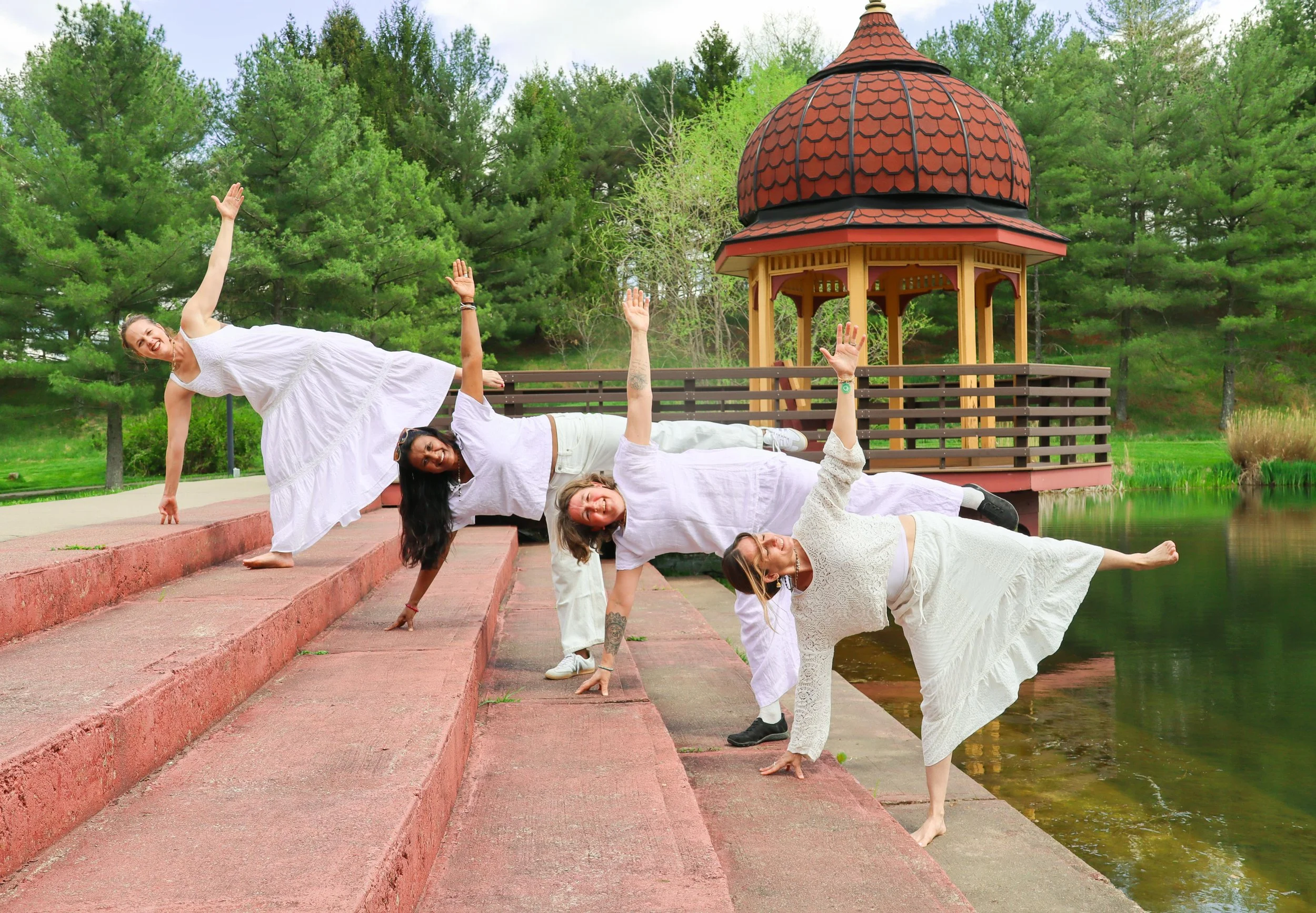 Five women in white dresses practicing yoga outdoors beside a pond with a gazebo and green trees in the background.