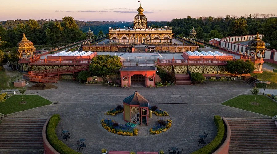 An ornate temple with golden domes, surrounded by lush trees and a spacious courtyard with garden beds and pathways.