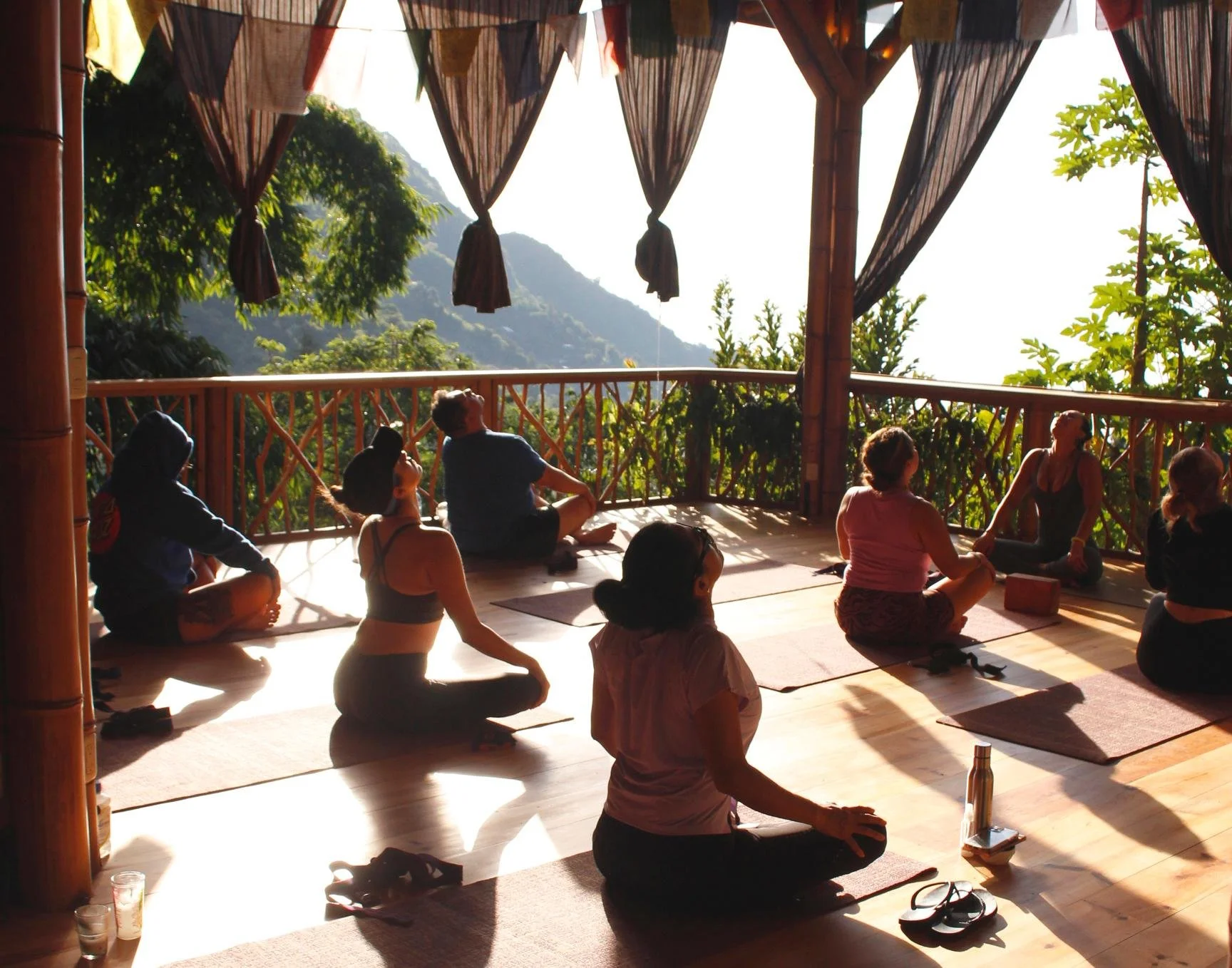 A group of people participating in a yoga or meditation class on a wooden deck overlooking a lush, green mountainous landscape during sunrise or sunset. They are seated on mats, facing outward, with some in a cross-legged position, practicing meditat
