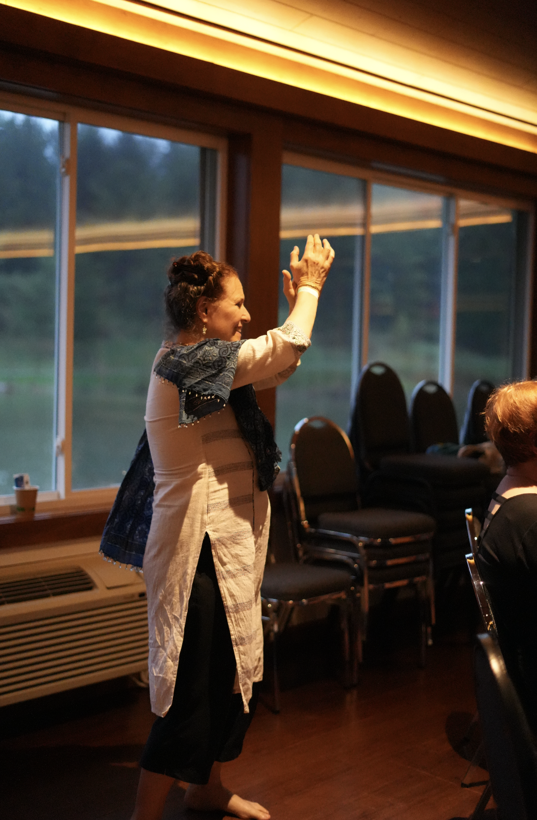 A woman in a beige shirt and black pants standing and smiling inside a room with large windows, clapping her hands, during an event or gathering.
