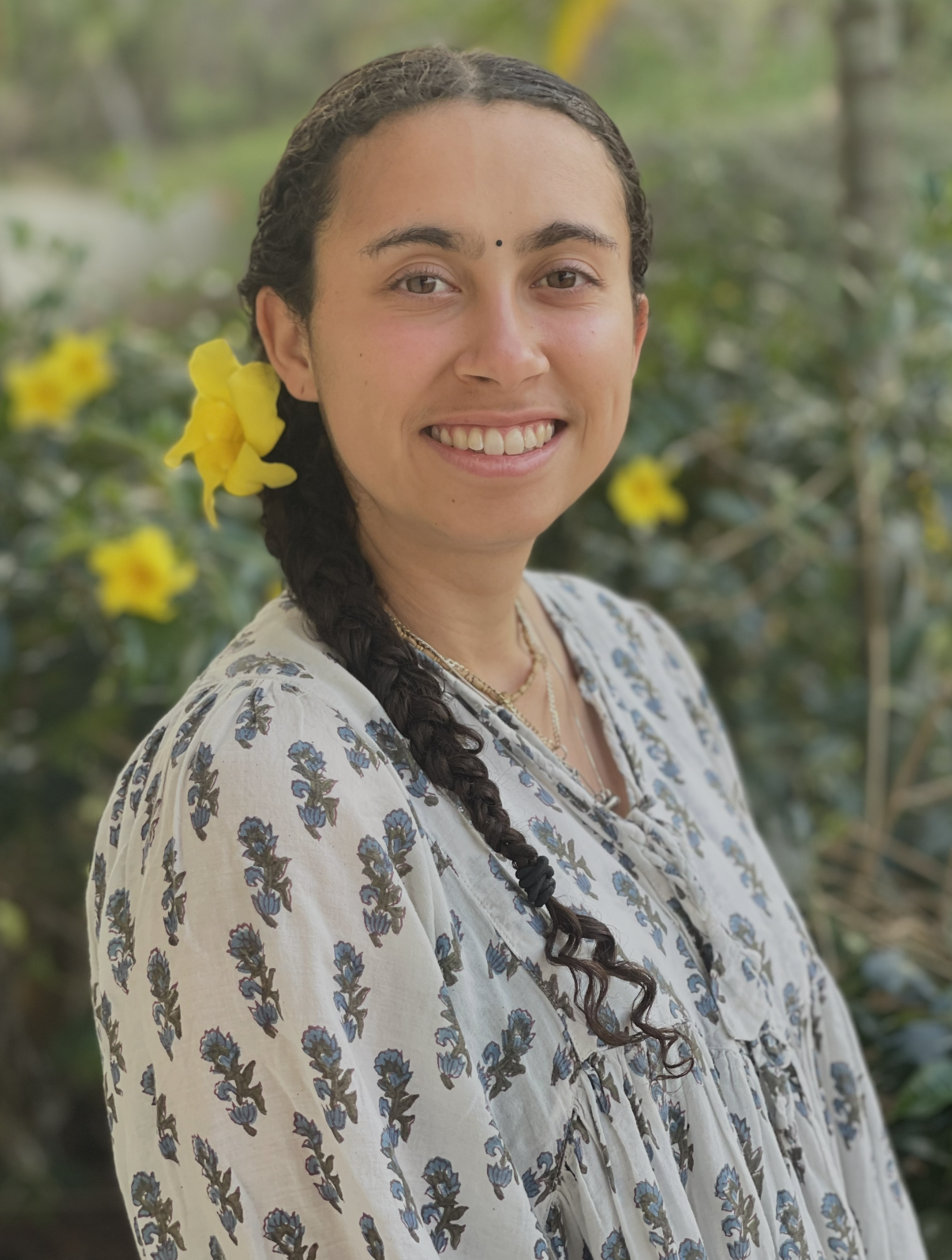 A woman with long braided dark hair, wearing a floral-patterned blouse and a small black bindi on her forehead, standing outdoors in a garden with yellow flowers and green foliage in the background.