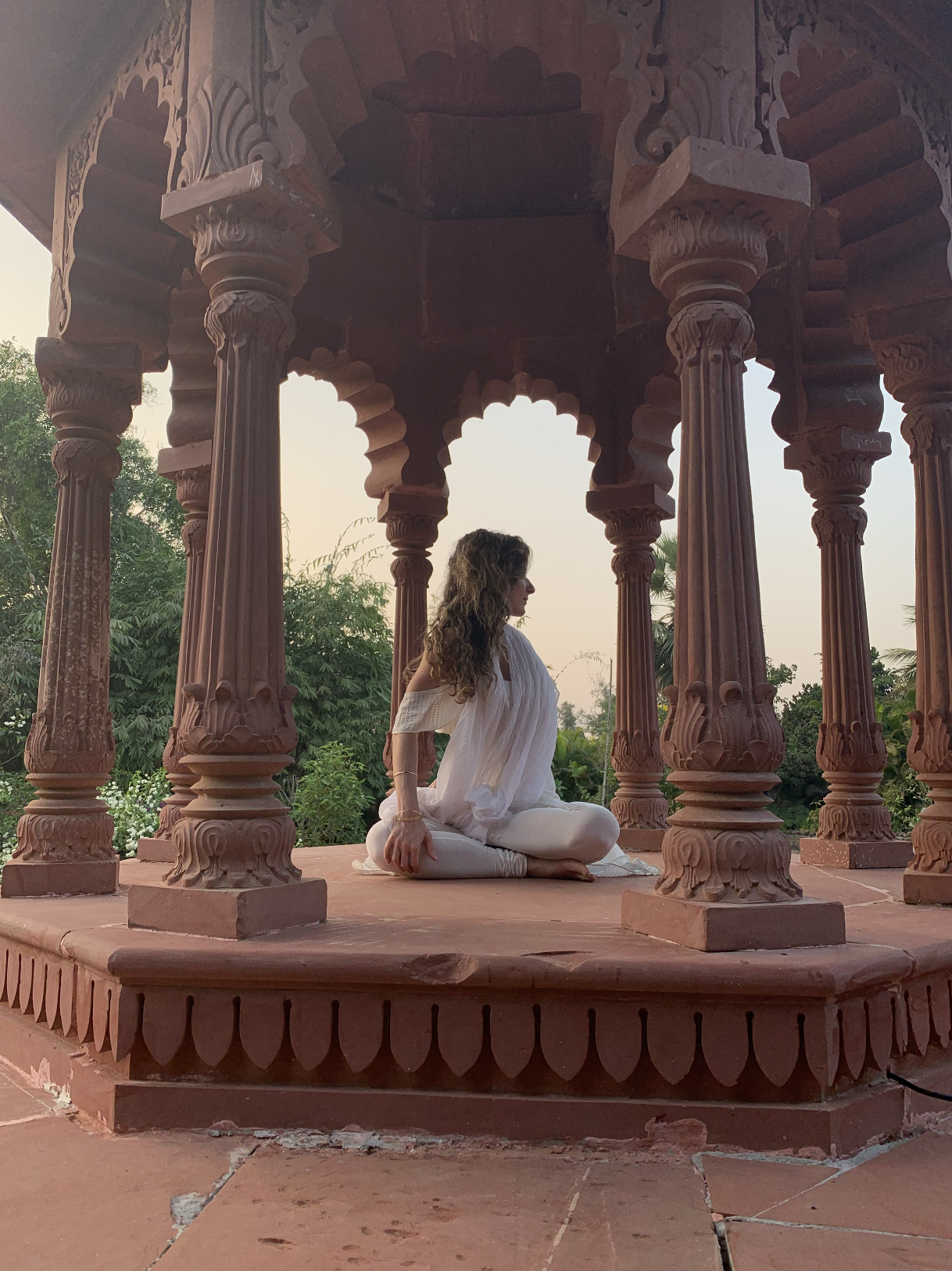 A woman with curly hair dressed in white, sitting cross-legged in a meditative pose inside a red sandstone pavilion with intricately carved pillars and an ornate ceiling, surrounded by greenery during sunset.