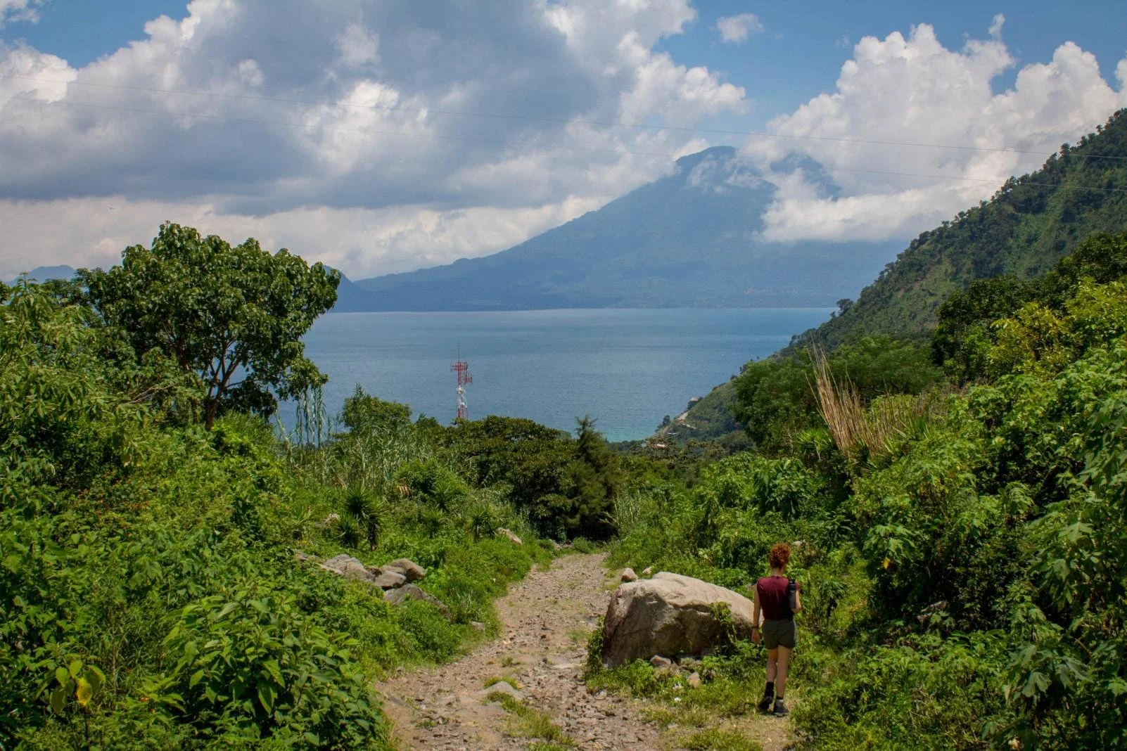 Woman hiking on a dirt trail through lush green vegetation towards a large body of water, with mountains and a partly cloudy sky in the background.
