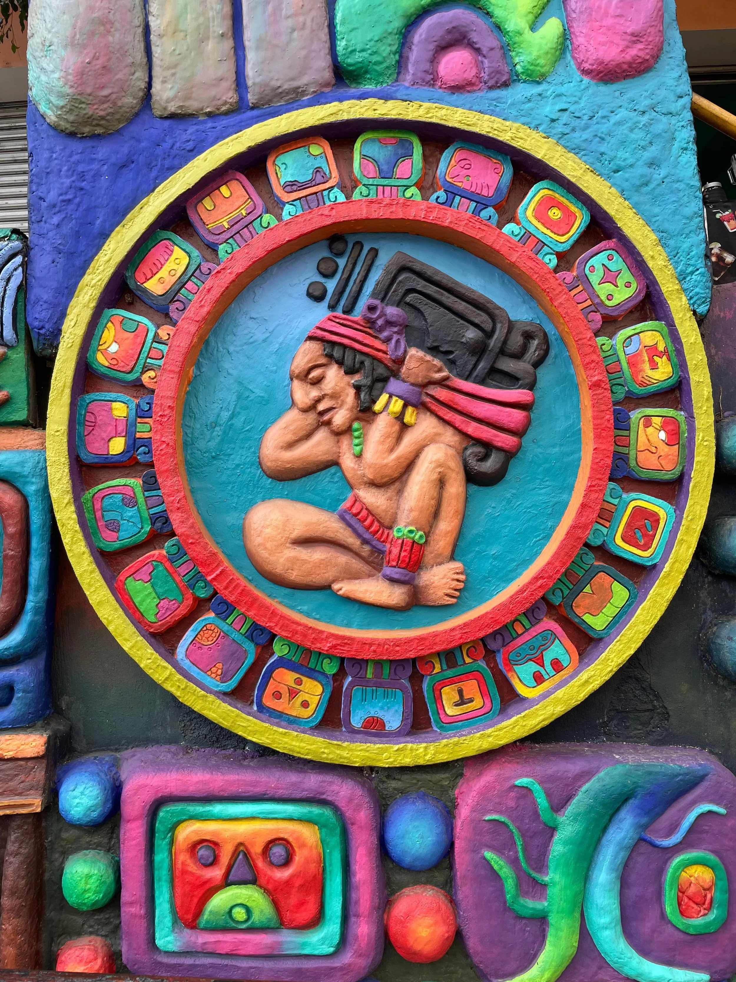 Colorful mural depicting a vibrant aztec calendar with a central figure, intricate designs, and bright colors including purple, yellow, green, red, and turquoise.