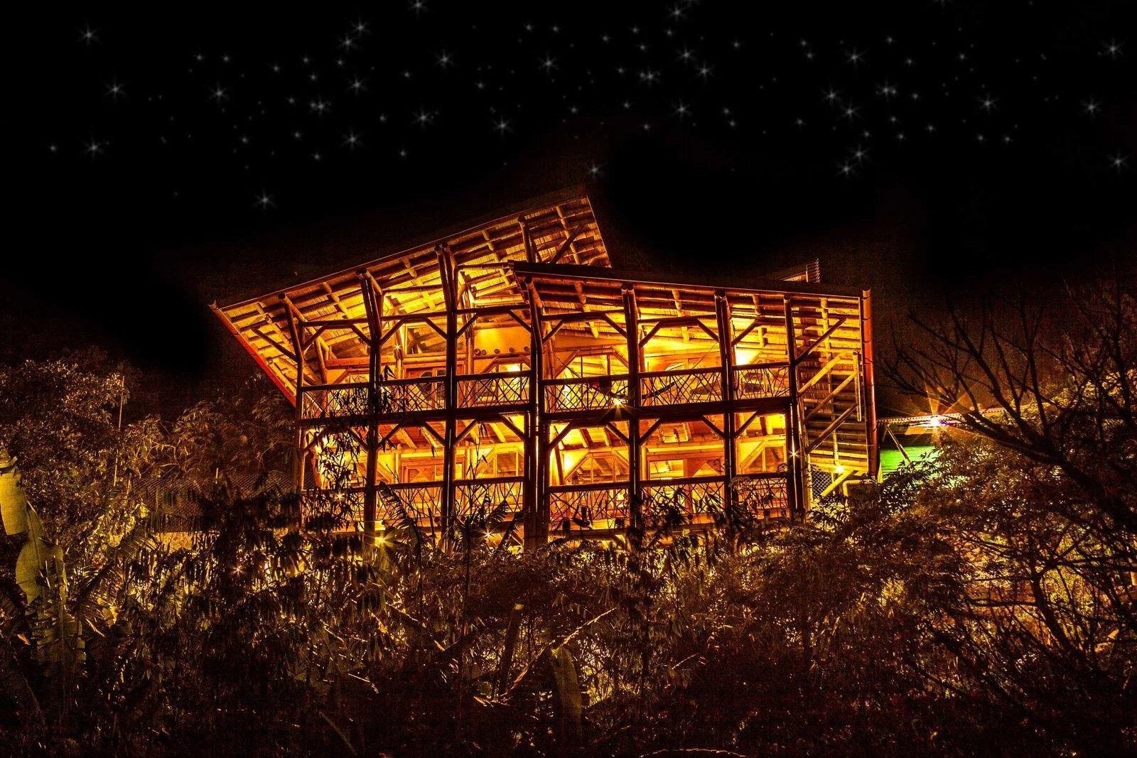 Nighttime view of a multi-level wooden structure, illuminated with orange lighting, surrounded by trees and bushes, with a starry sky in the background.