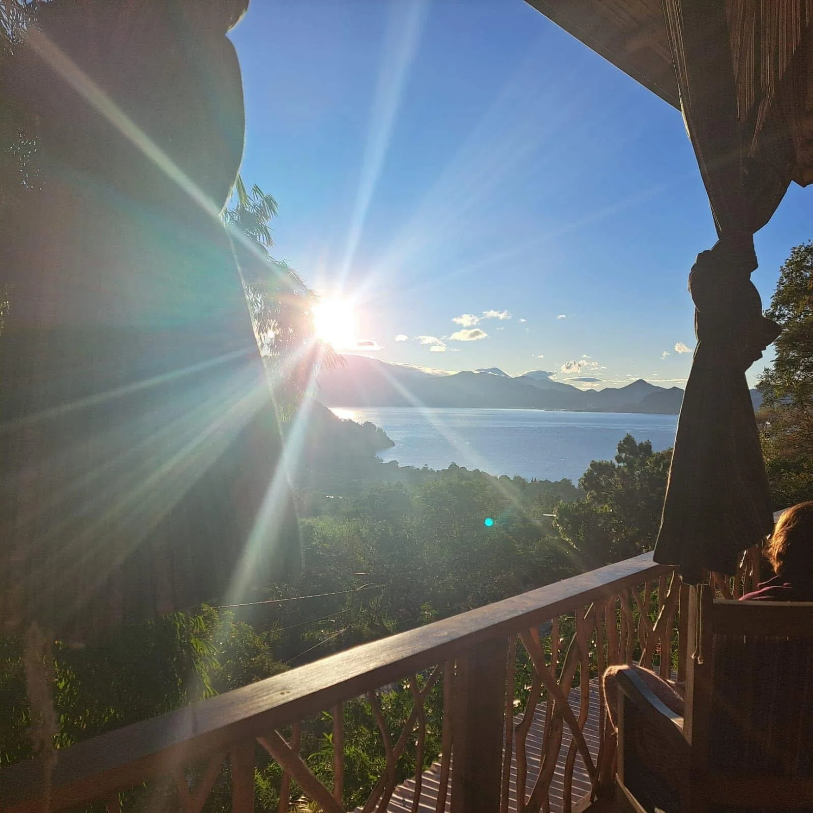 Sunset over a lake viewed from a wooden balcony with trees and mountains in the background.