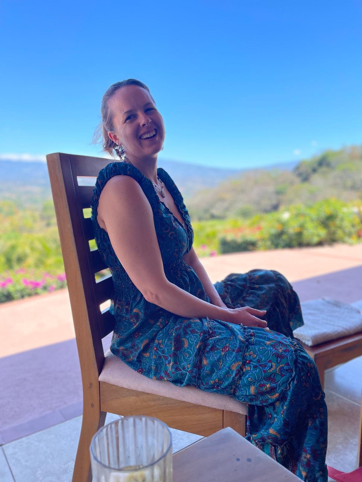 A woman wearing a blue patterned dress is sitting on a wooden chair outdoors with a scenic mountain and greenery background. She is smiling and looking at the camera.