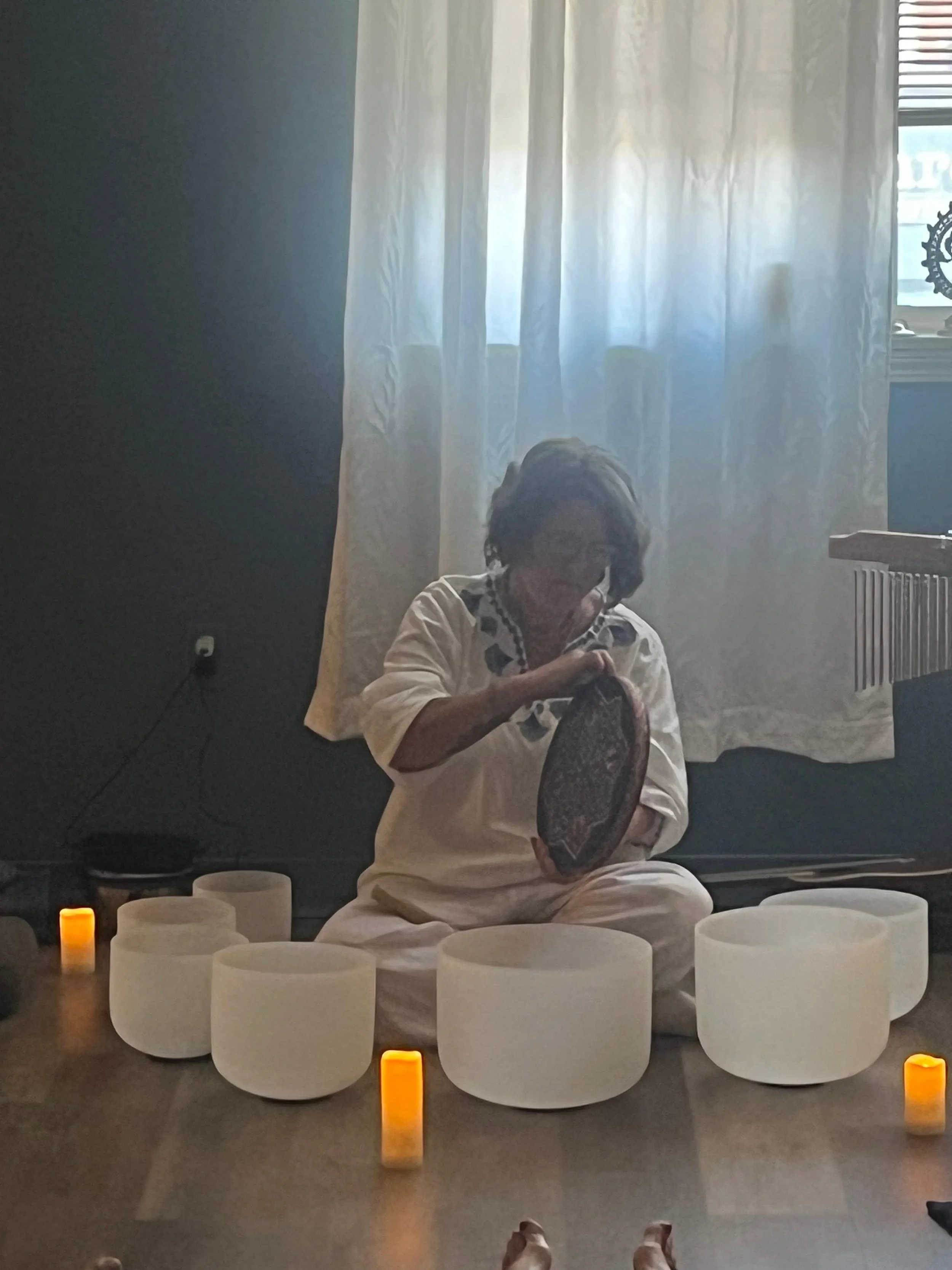 A woman seated on the floor wearing a white outfit, holding a drum and facing a display of candles and white cylindrical objects, with a backdrop of a sheer curtain and a window with partially closed blinds.