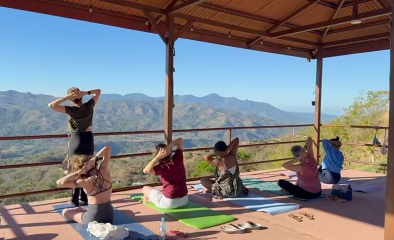 Group of women practicing yoga outdoors on a wooden platform with mountain view, sitting on mats and performing a seated stretch with hands behind heads under a roofed shelter.
