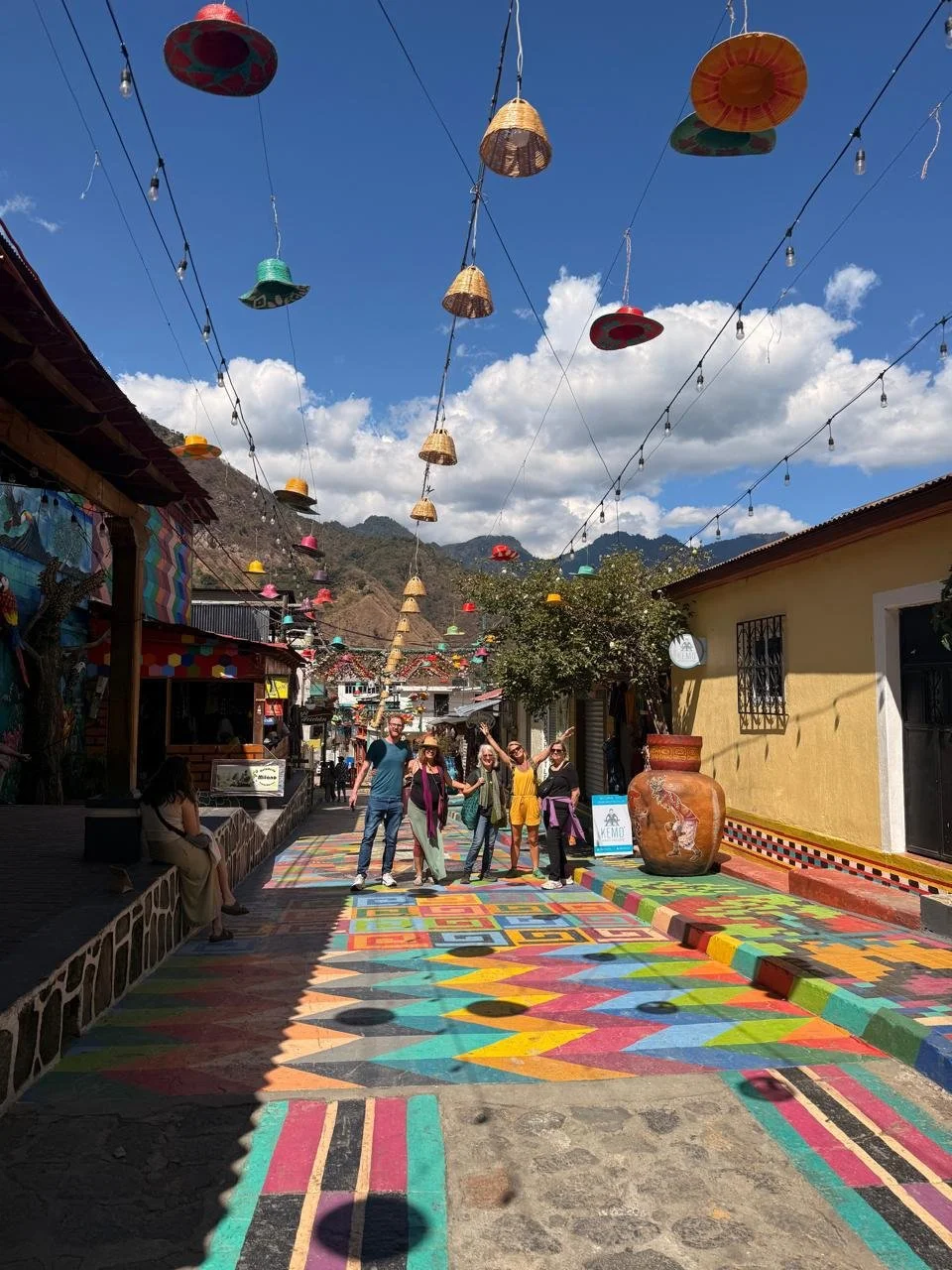 Colorful decorated street with hanging hats and string lights, with a group of people posing and taking photos under a cloudy sky in a town with mountains in the background.