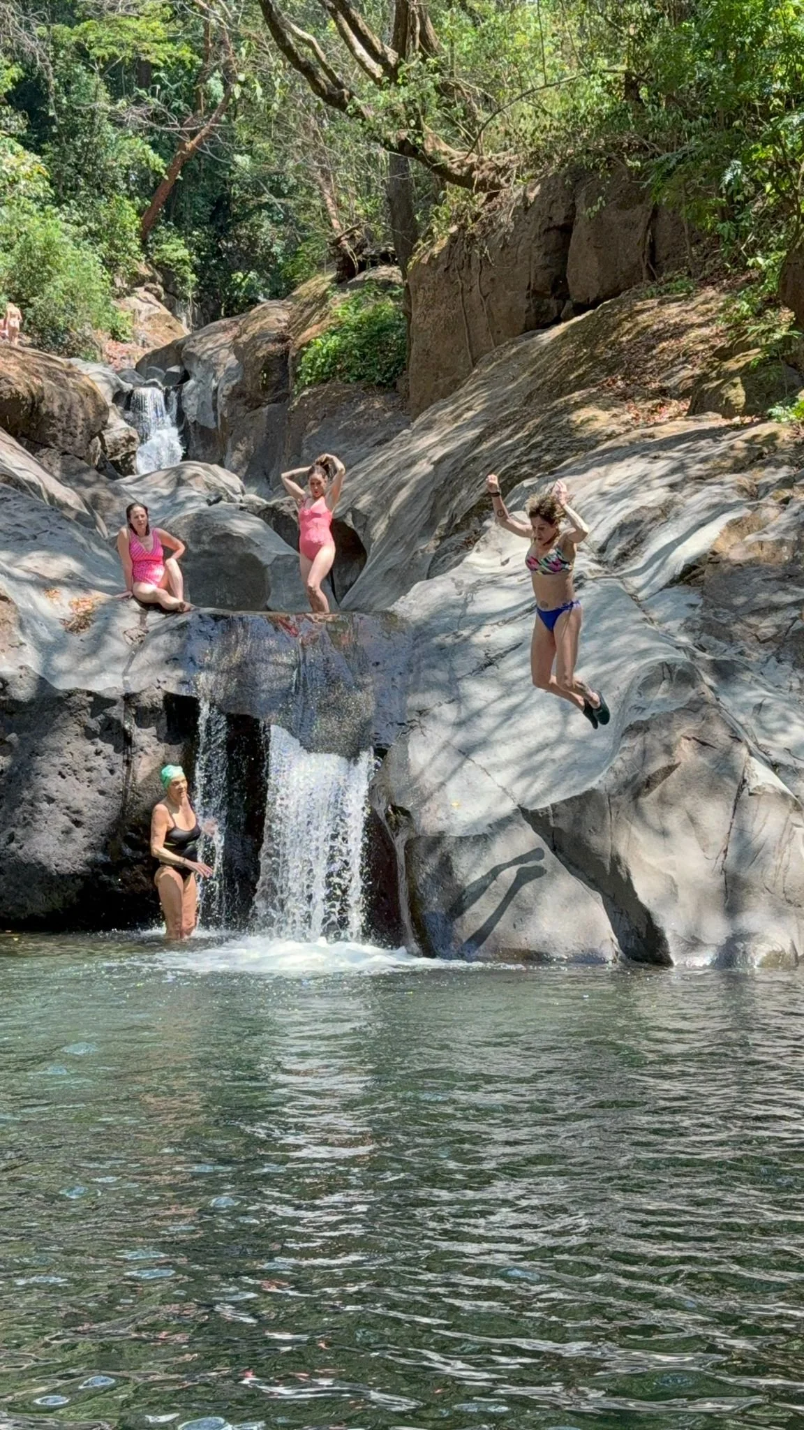 Four women enjoying a day by a rocky waterfall and swimming hole, some sitting on rocks, one jumping off a rock, and another standing in the water, surrounded by lush green trees.