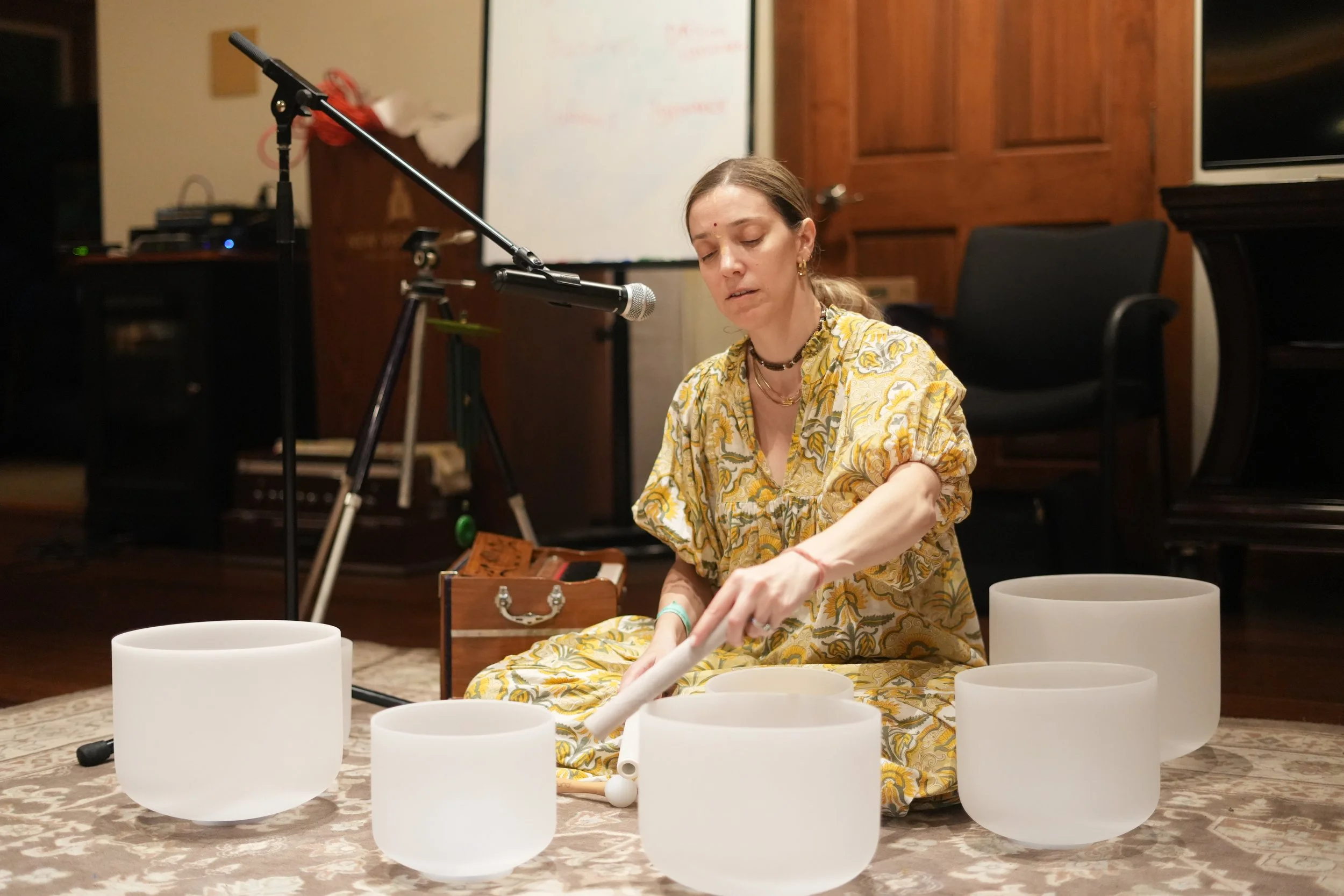 A woman with light brown hair and wearing a yellow patterned dress is sitting on the floor, playing crystal singing bowls. There are multiple crystal bowls in front of her and she is using a mallet to play one. In the background, there is a whiteboar