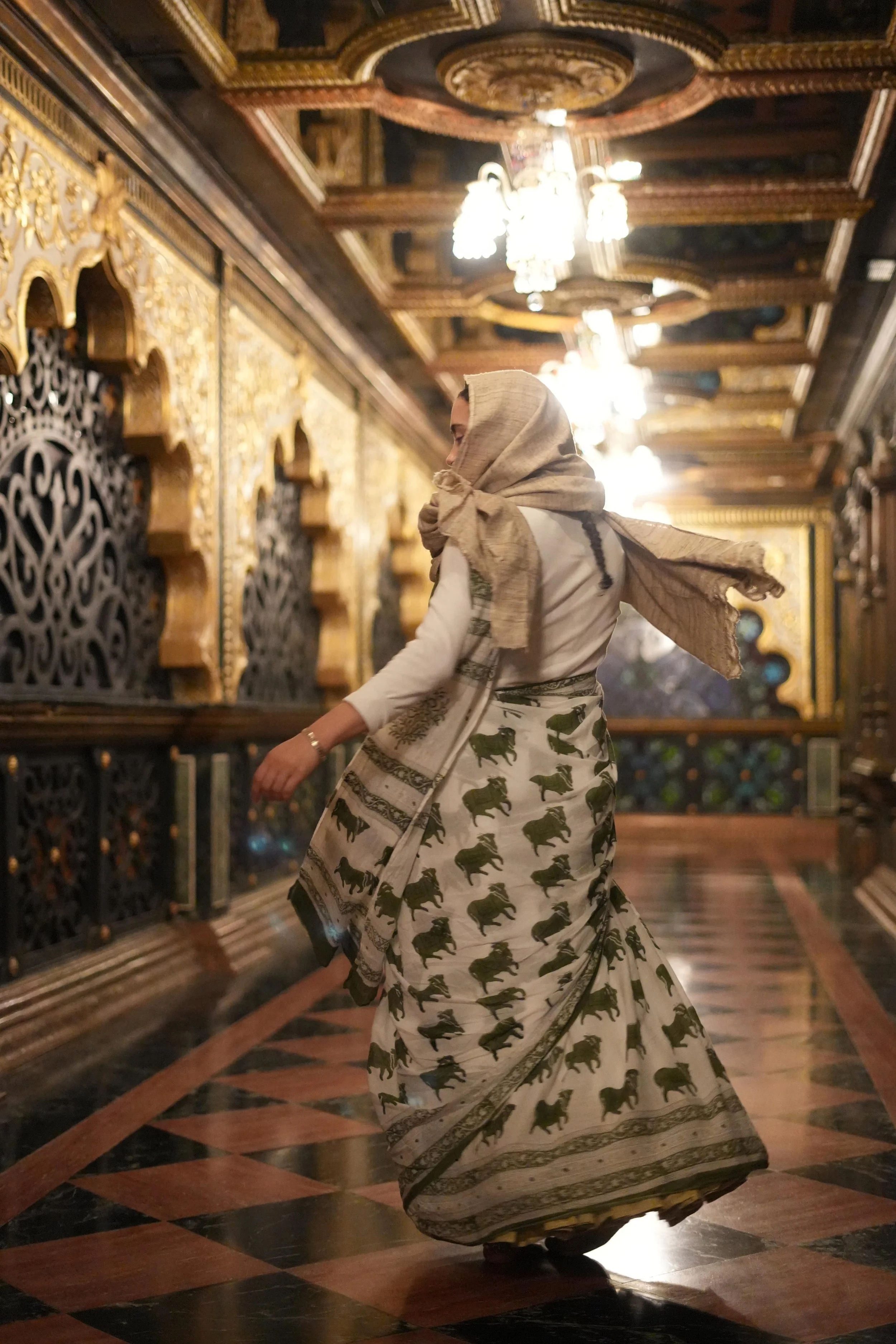 A woman wearing a beige headscarf and a traditional Indian saree with elephant motifs is spinning or dancing in an ornately decorated room with intricate woodwork and chandeliers.