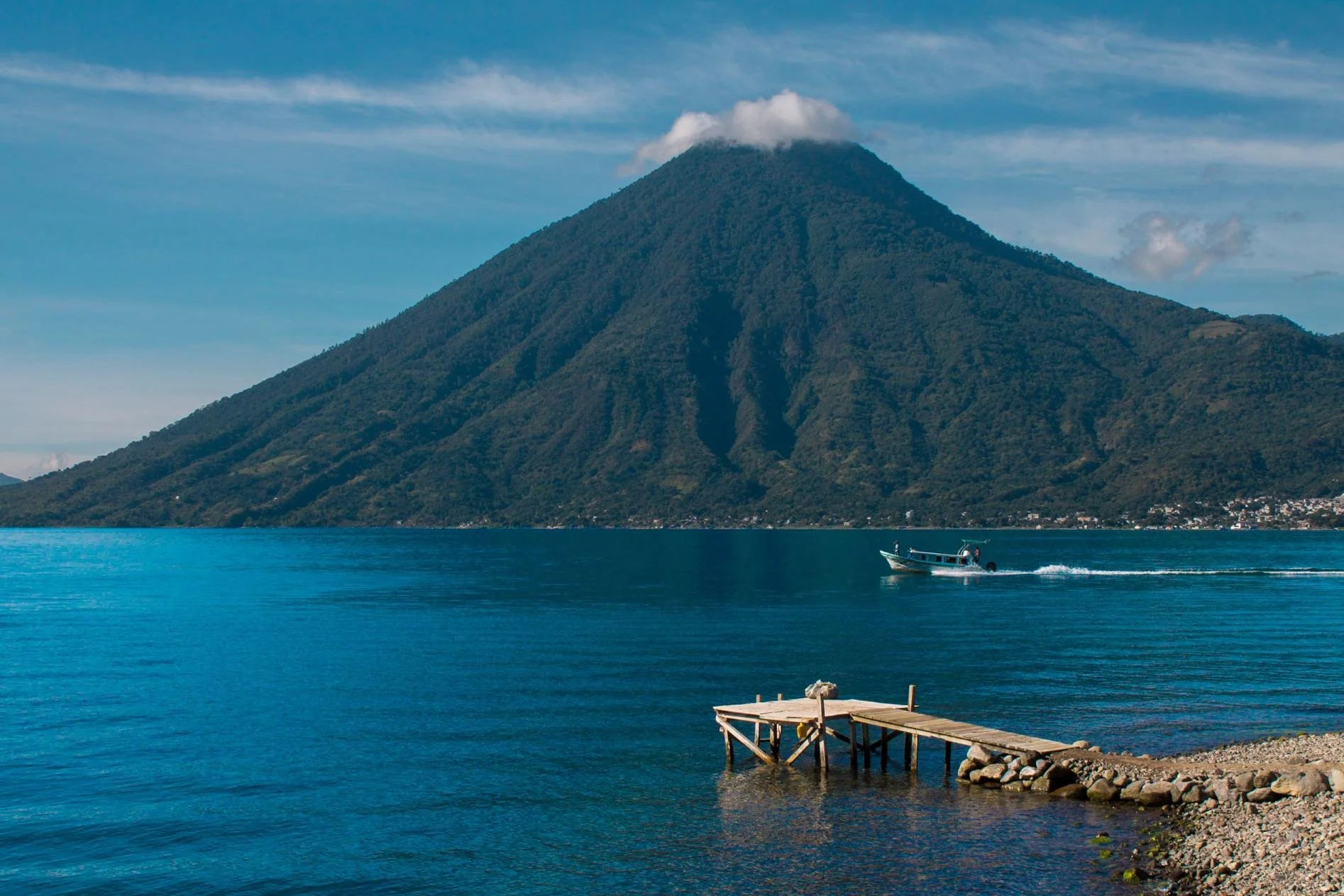 A boat sailing on a large blue body of water with a green, cone-shaped mountain with a small cloud on top in the background and a wooden dock in the foreground.